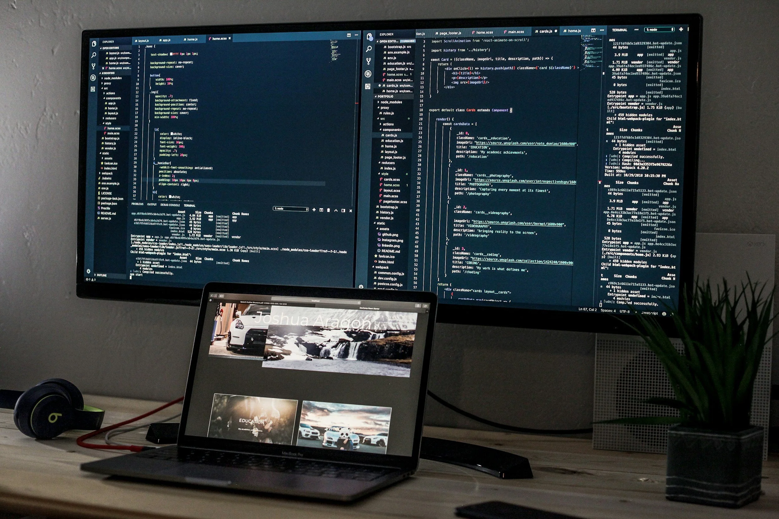 A desk setup with a large monitor displaying lines of code, a laptop showing a photo gallery, a pair of Beats headphones, and a potted plant on the right side.