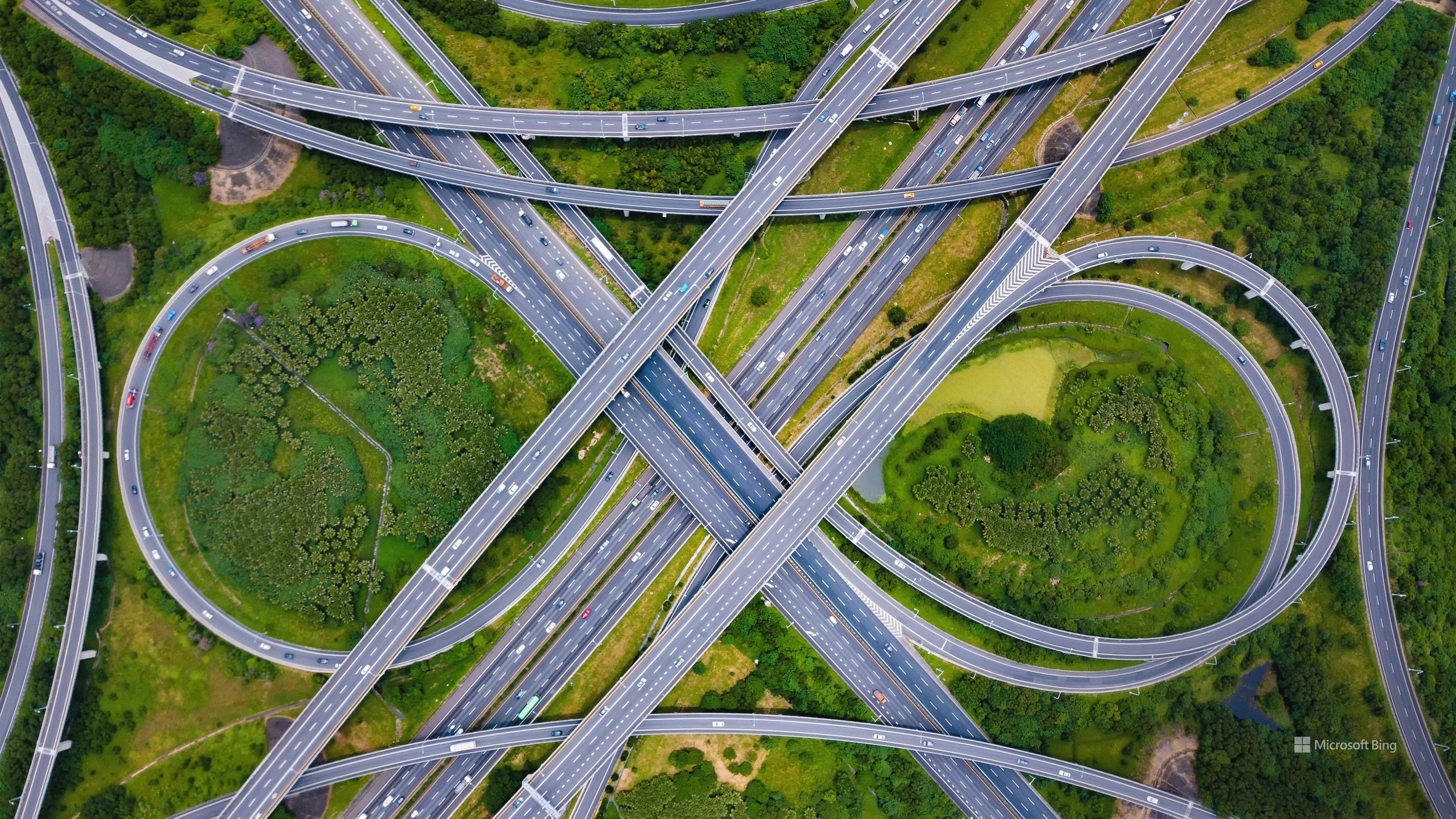 Aerial view of a complex highway interchange with multiple looping ramps and overpasses surrounded by green trees and grass.