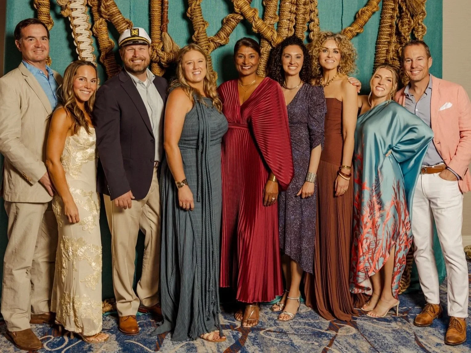 Group of people dressed in formal attire, standing in front of a nautical-themed backdrop with ropes, smiling at the camera.