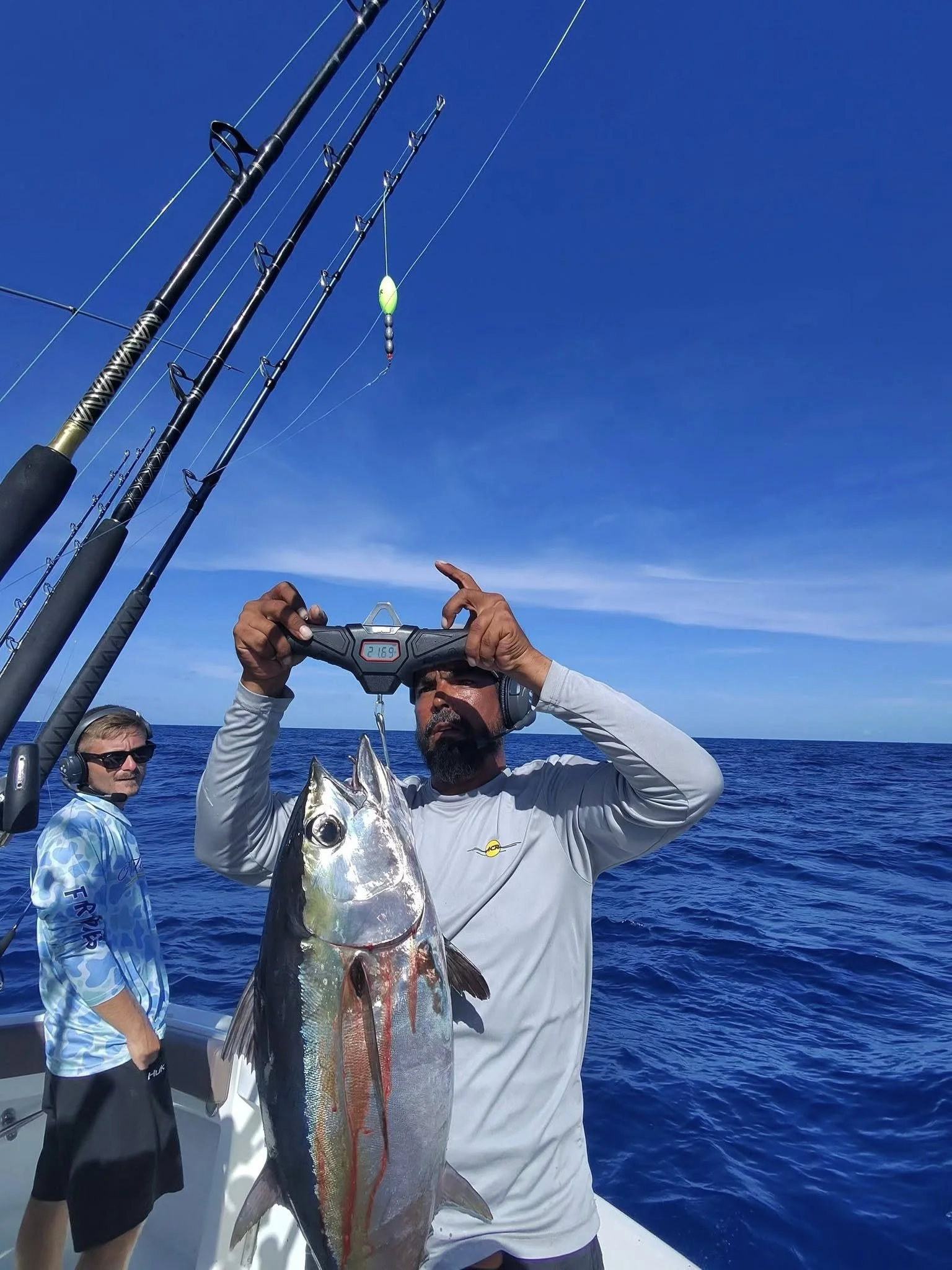 A man holding a large fish he caught with a digital scale showing 21.69 pounds on a fishing trip at sea, with two fishing poles and clear blue sky in the background.