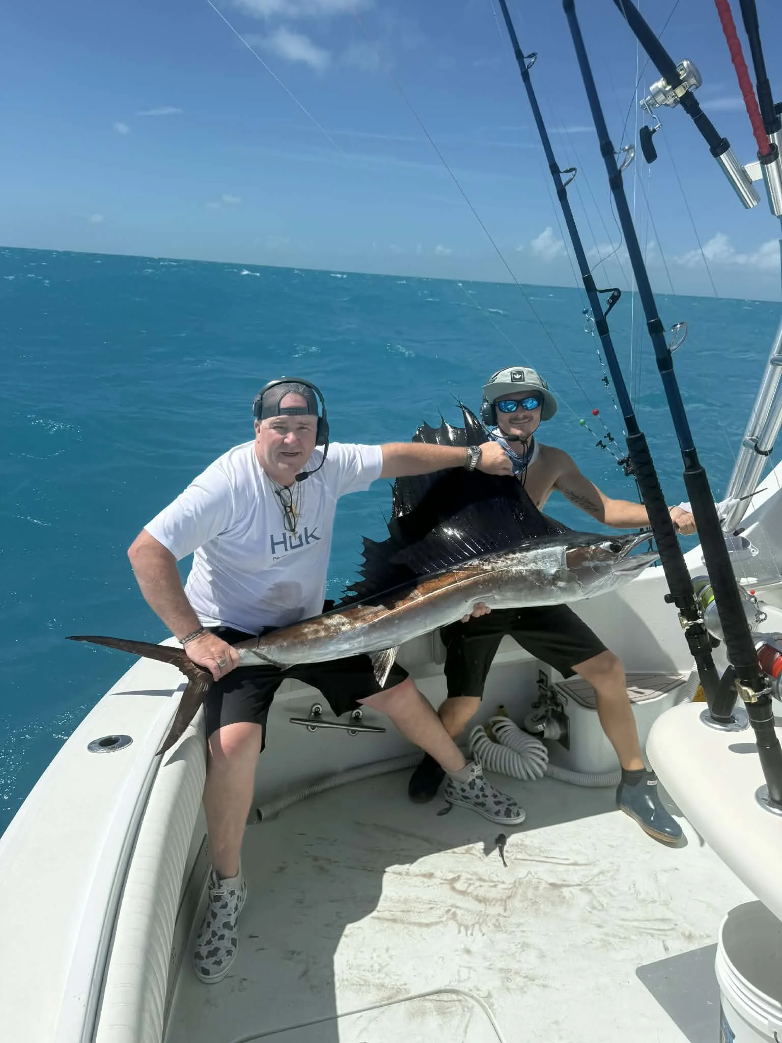 Two men on a boat holding a large fish they caught, with fishing rods and ocean in the background.