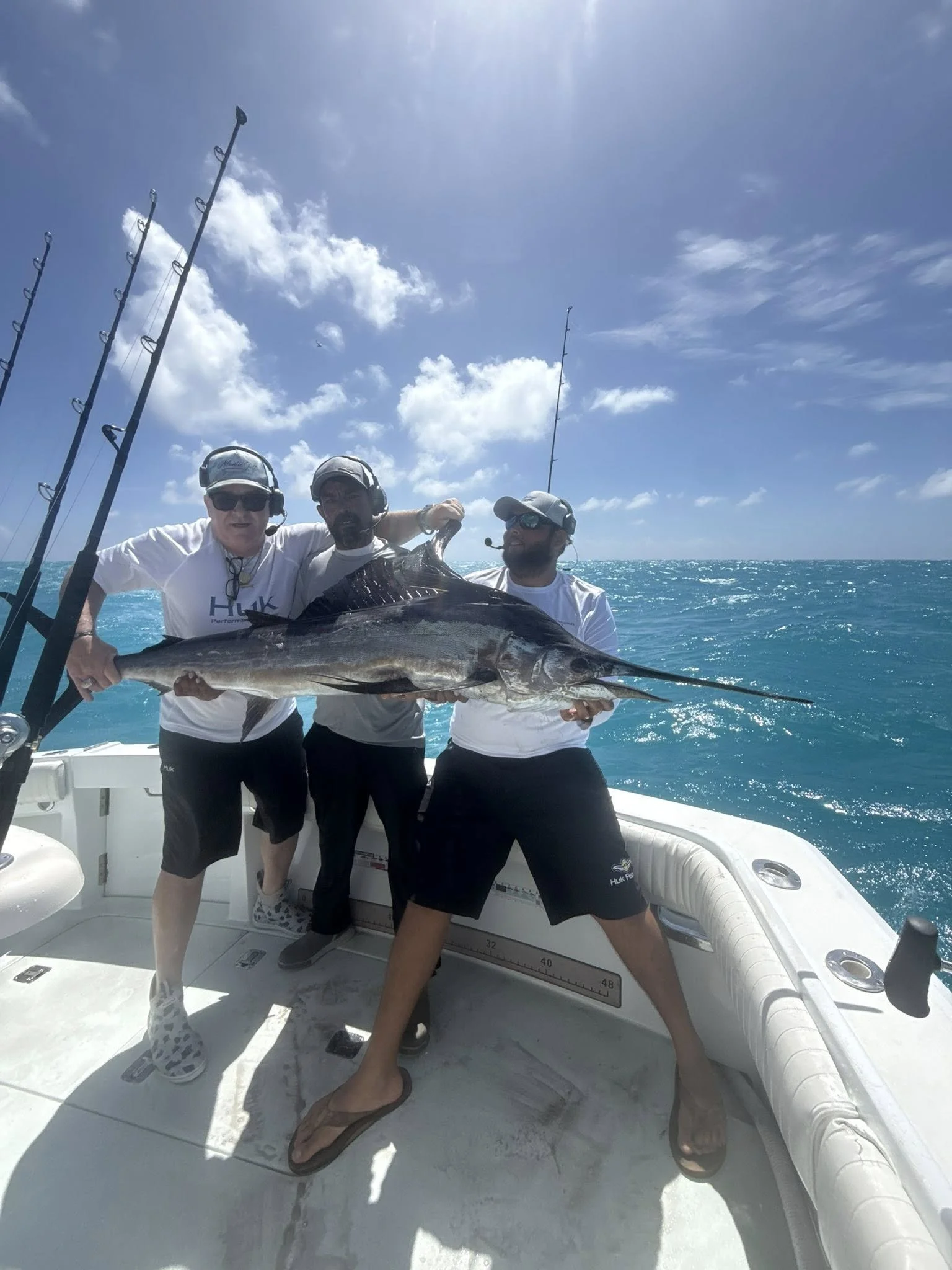 Three men on a boat holding a large fish they caught, with fishing rods on the side and ocean in the background.