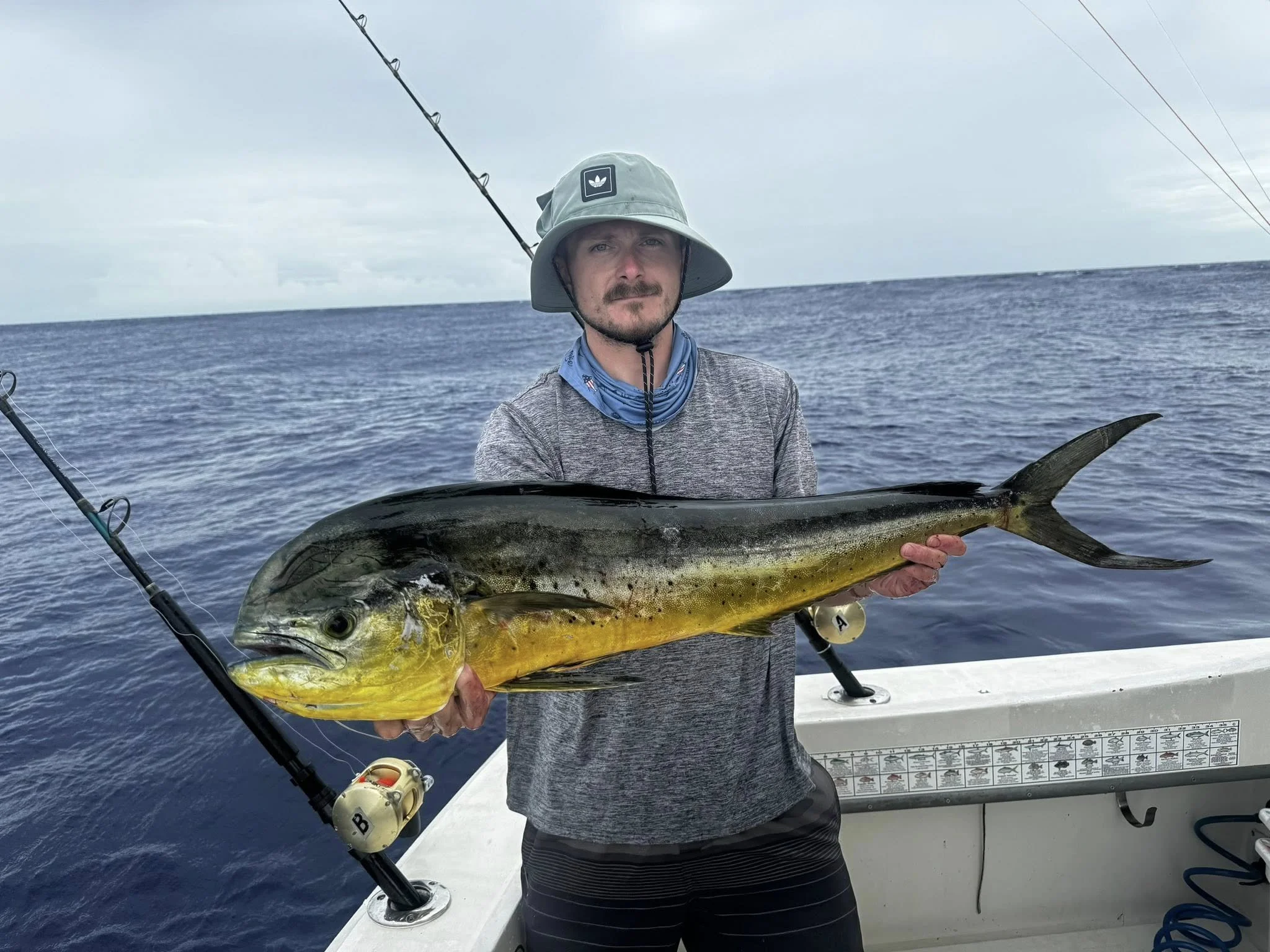 A man on a boat holding a large fish, with fishing rods in the background and the open sea behind him.