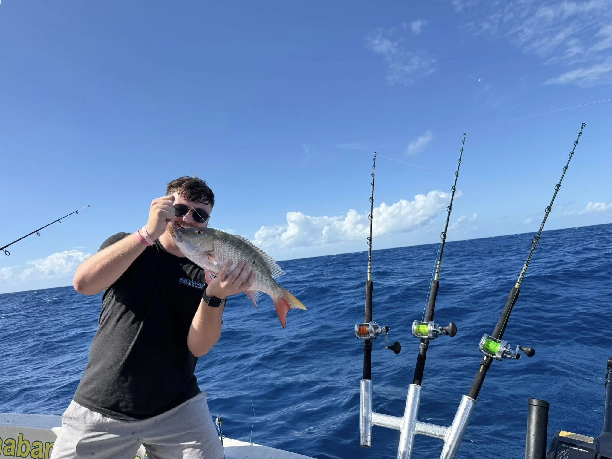 A man wearing sunglasses and a black t-shirt holding a caught fish on a boat with fishing rods in the ocean under a blue sky with clouds.