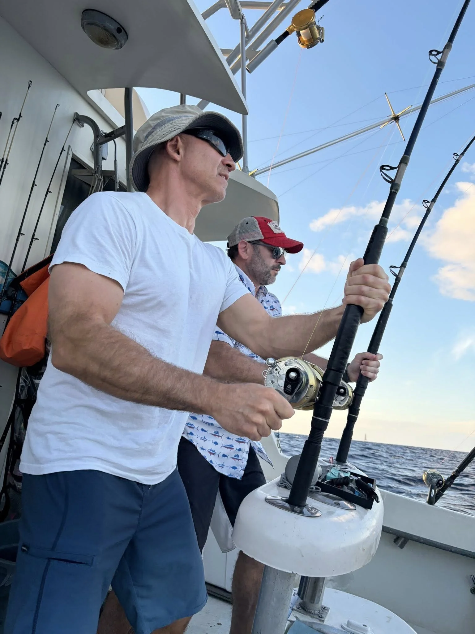 Two men fishing on a boat at sea, one wearing a white t-shirt, wide-brim hat, sunglasses, and holding a fishing rod, the other wearing a patterned shirt, red cap, and sunglasses, also holding a fishing rod.