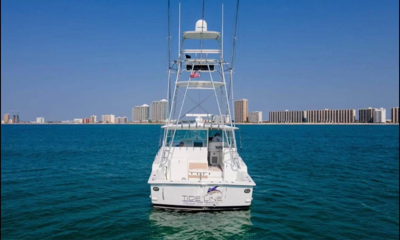 White fishing boat on blue ocean waters with a city skyline of high-rise buildings in the background under a clear blue sky.