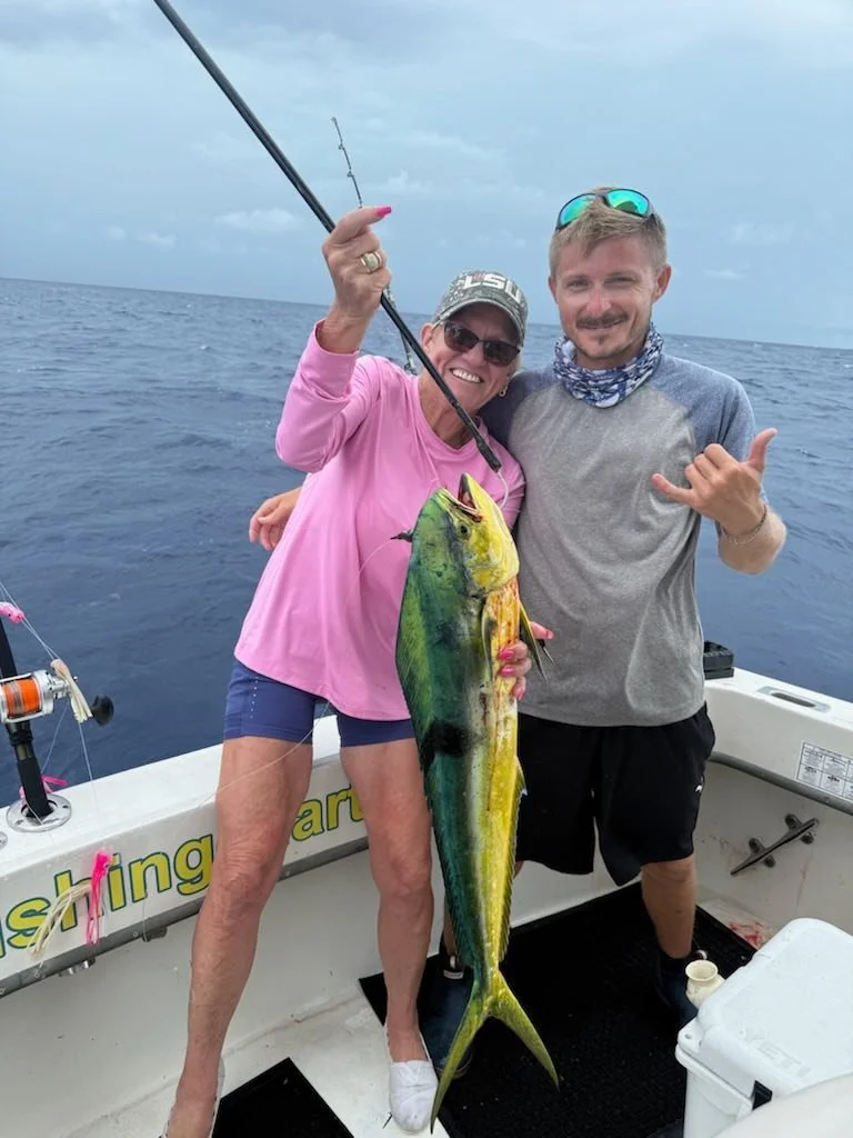 Two people on a boat at sea, holding a large fish they caught. The woman on the left is wearing a pink shirt, shorts, and sunglasses, holding the fishing rod and smiling. The man on the right is wearing a gray shirt, shorts, sunglasses on his head, and a neck gaiter, making a shaka hand sign.