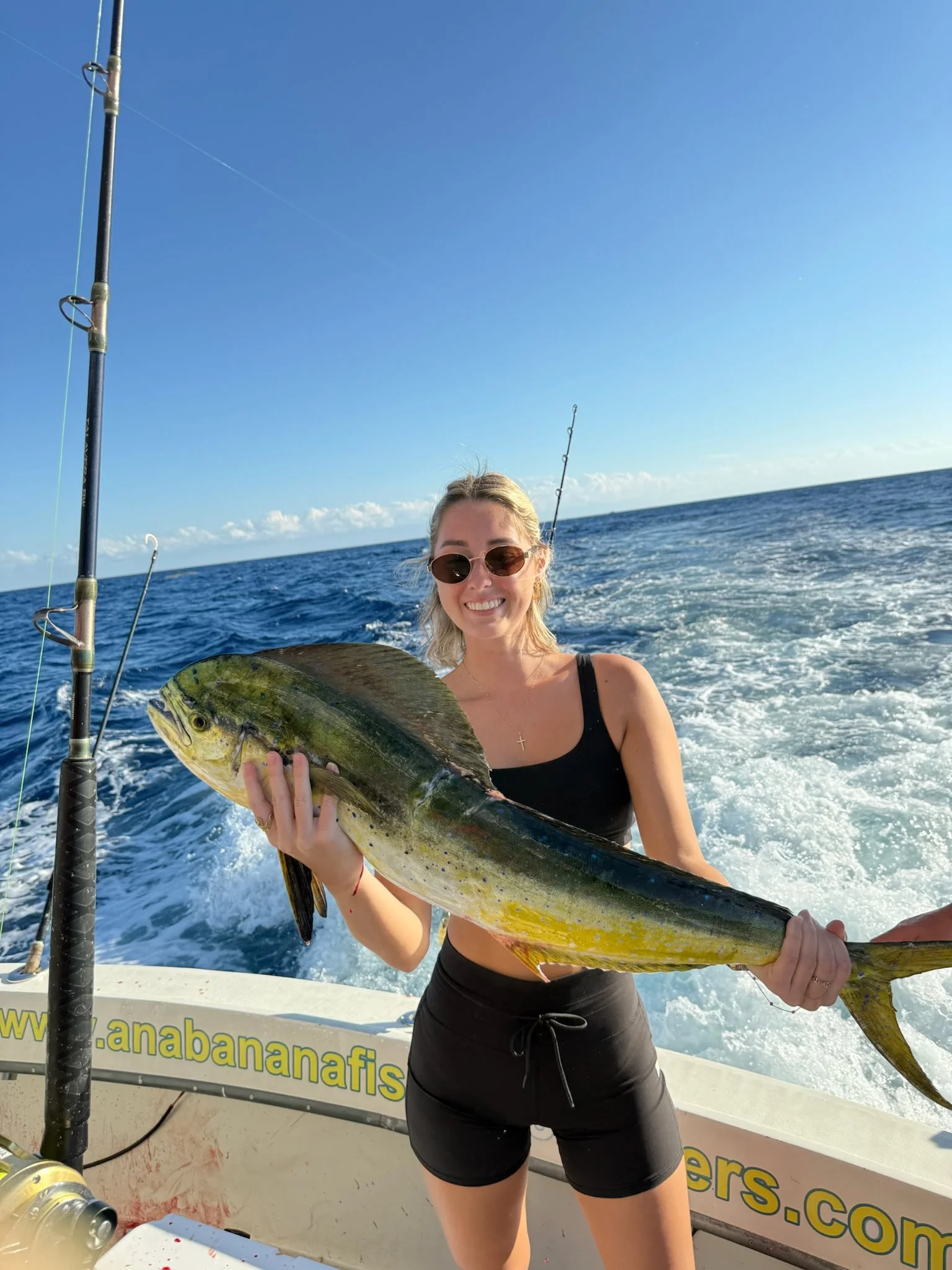 A woman wearing sunglasses and a black tank top holding a large fish on a boat in the ocean, with fishing rods visible and a clear blue sky in the background.