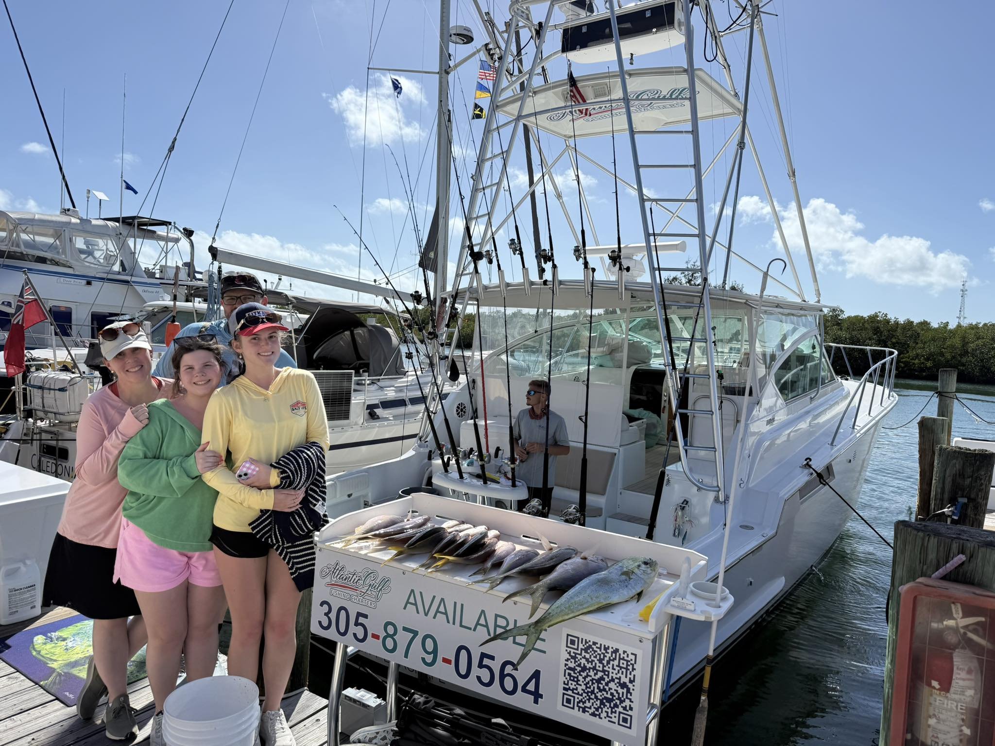 Four women standing on a dock next to a boat with fish laid out in front and a man fishing on the boat. The women are smiling at the camera, and the boat has fishing rods and a canopy. The dock has a QR code and phone number for rental or sales.