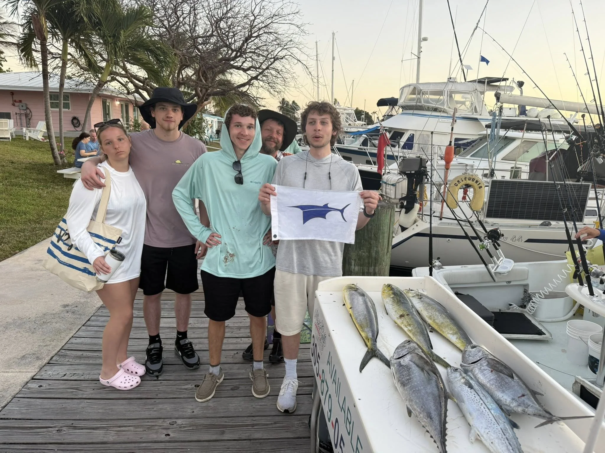 Group of six people standing on a dock with boats in the background, posing with a caught fish and a flag, with a tray of freshly caught fish in the foreground.