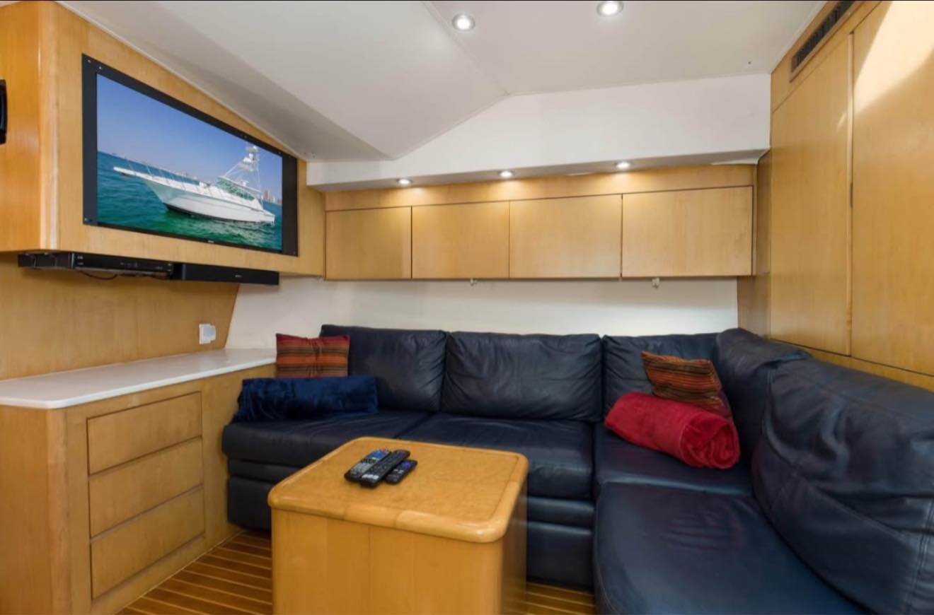 Interior of a boat or yacht living room with a black leather sectional sofa, wooden cabinetry, a small wooden table with TV remote controls, a flat-screen TV displaying a boat, and a few pillows on the sofa.