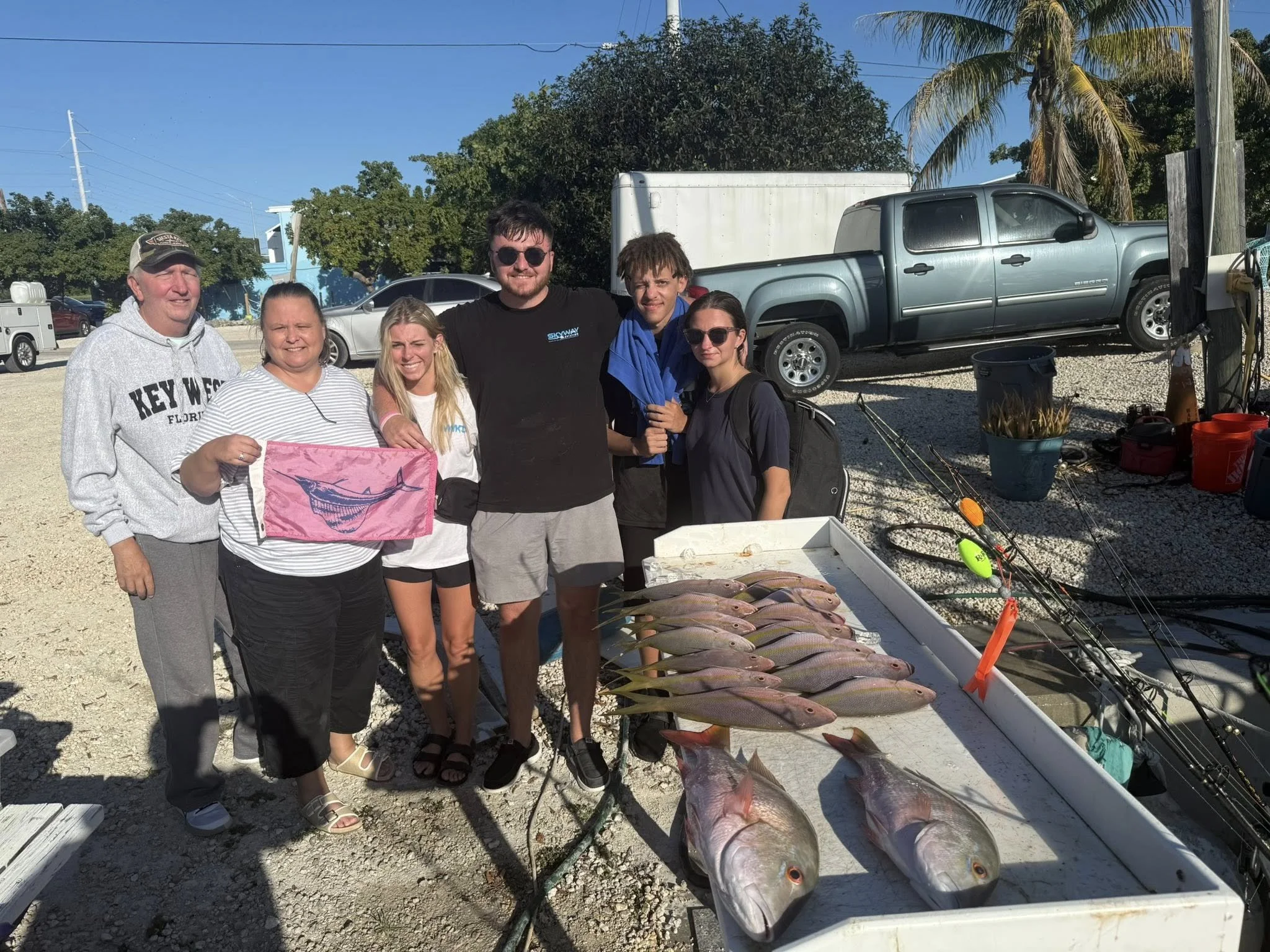 Group of six people standing behind a table of freshly caught fish and fishing gear outdoors on a sunny day.