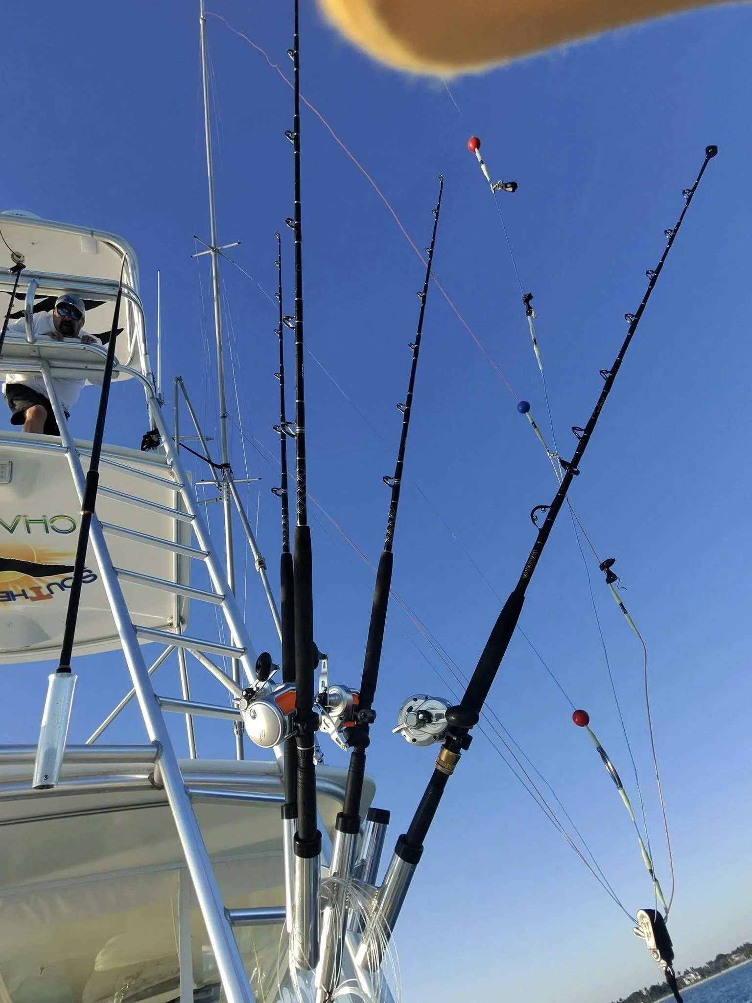 Four fishing rods secured to a boat's railing with a man on the boat holding on, set against a clear blue sky.