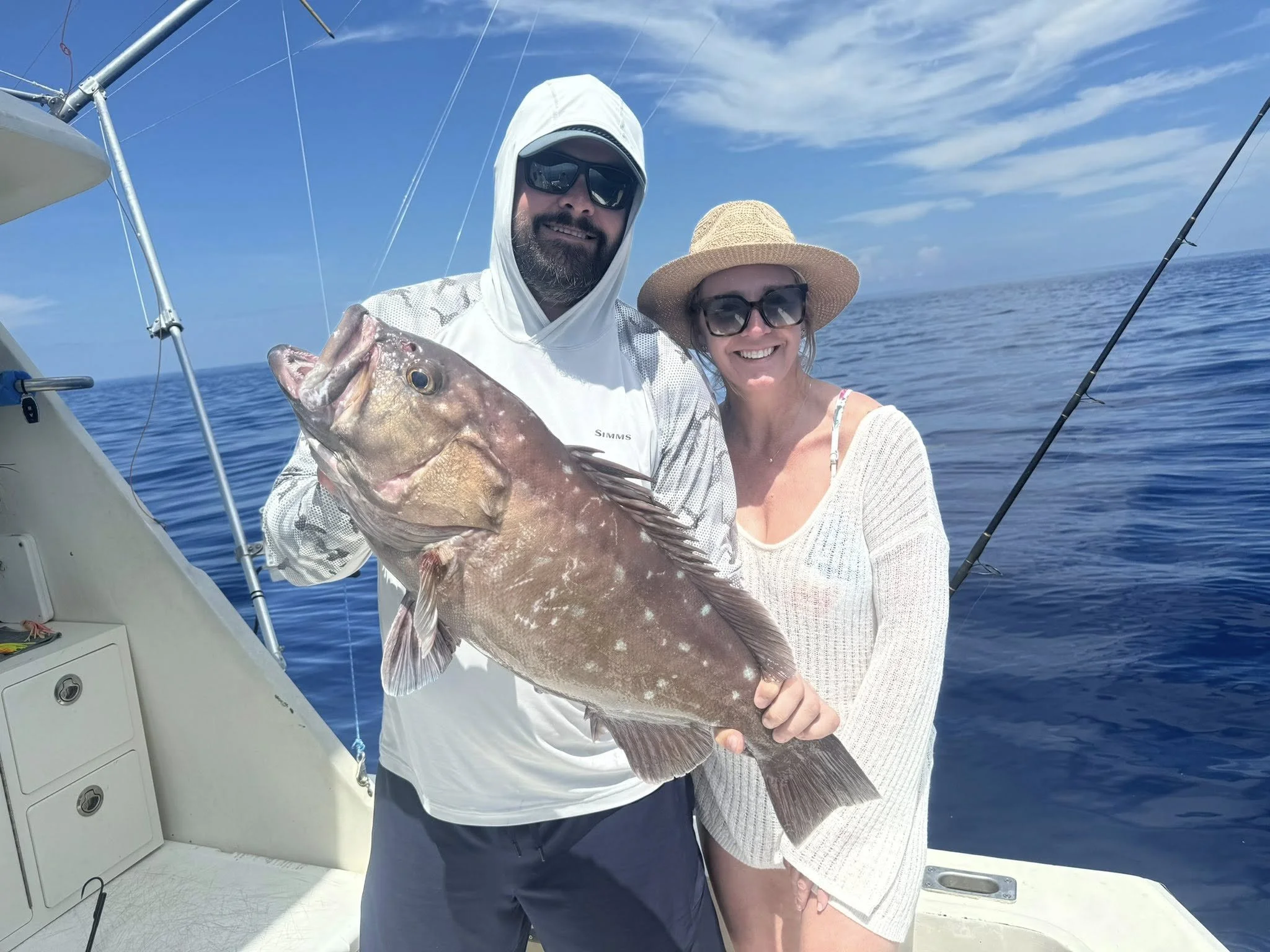 A man and a woman on a boat holding a large fish they caught, with blue ocean and sky in the background.