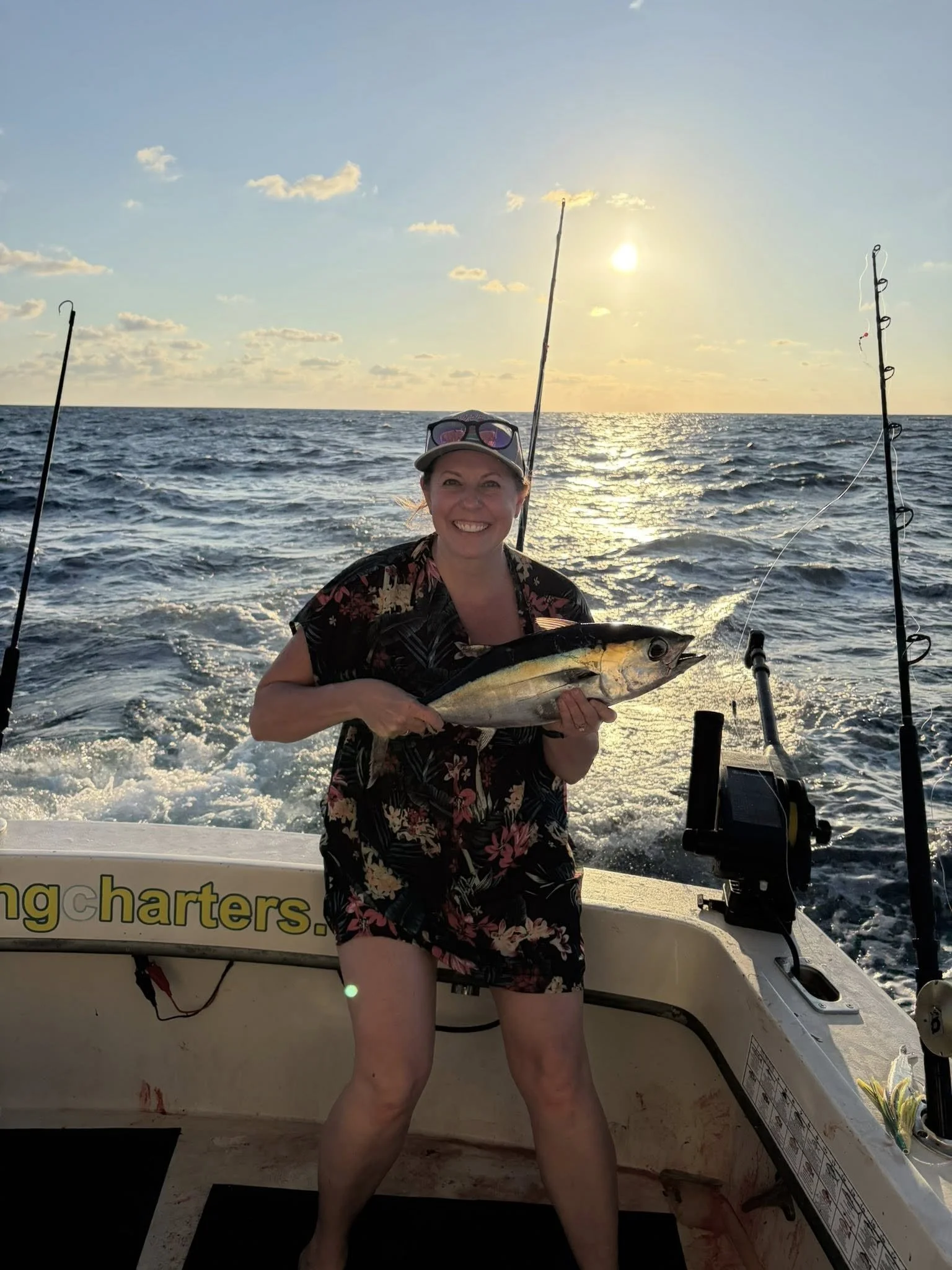 A woman smiling and holding a large fish on a boat at sunset, with fishing rods and the ocean in the background.