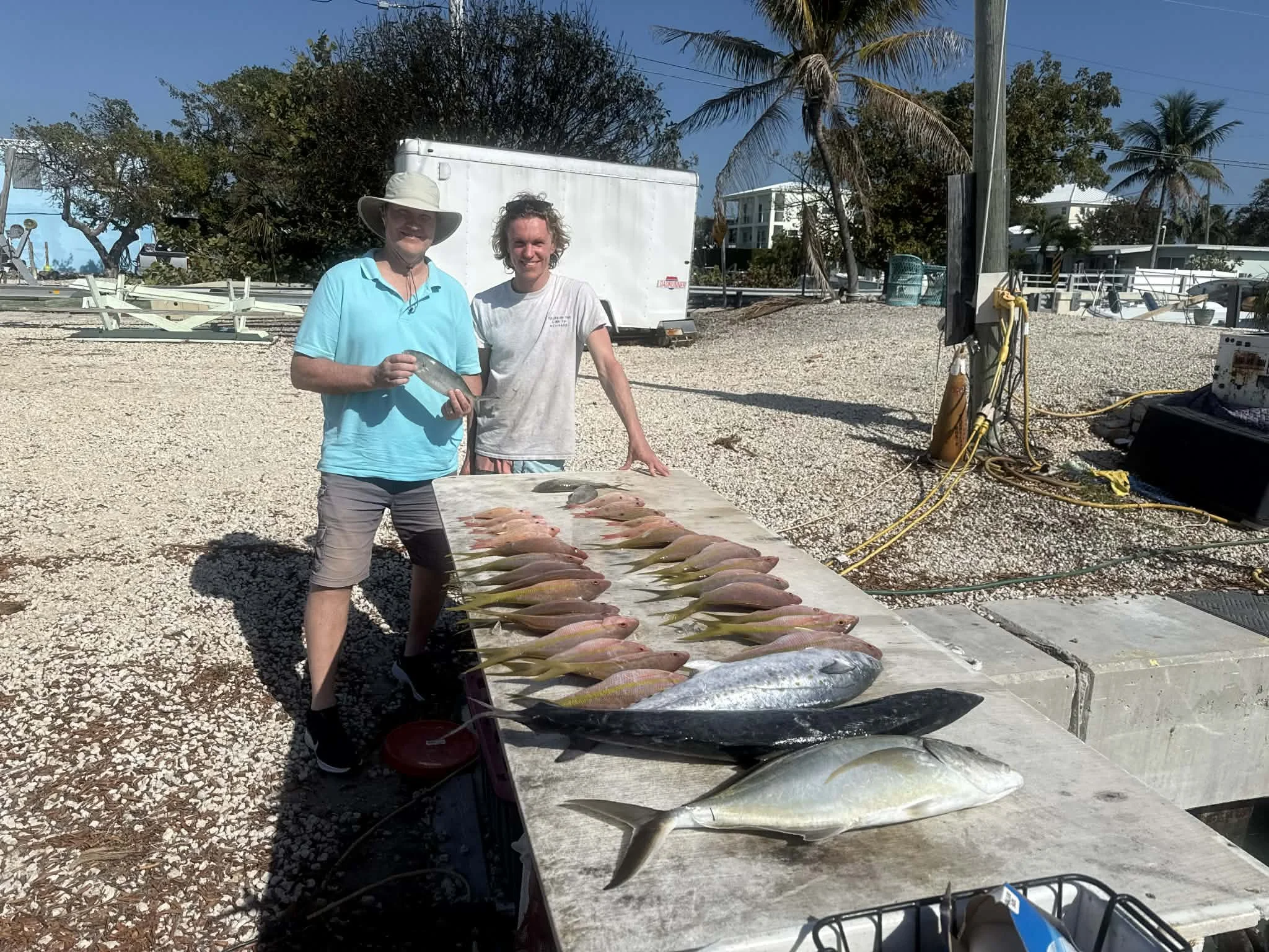Two men standing next to a table with freshly caught fish, at a marina or fishing dock on a sunny day.