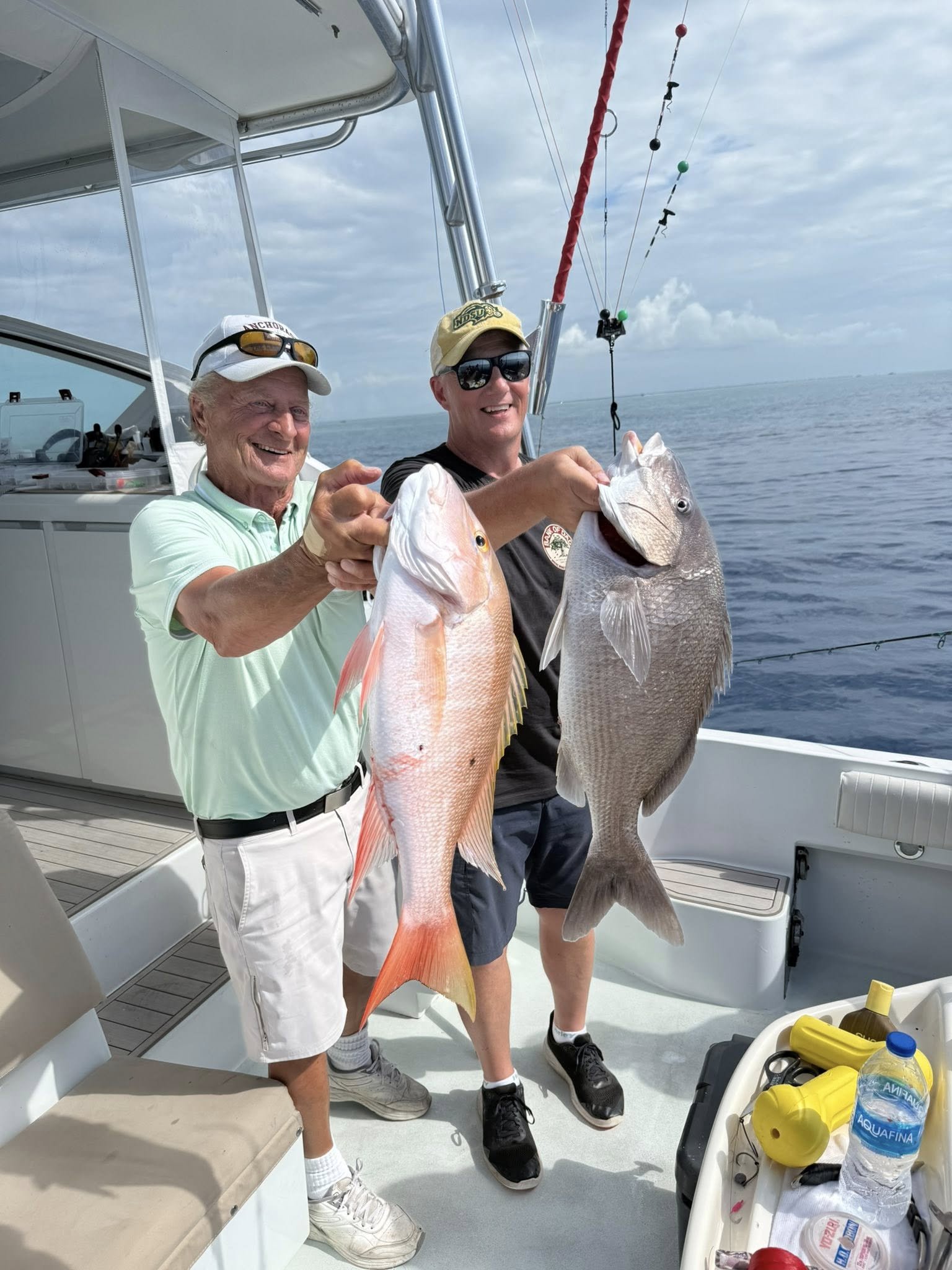 Two men smiling on a boat holding large fish they caught during a fishing trip, with ocean and cloudy sky in the background.