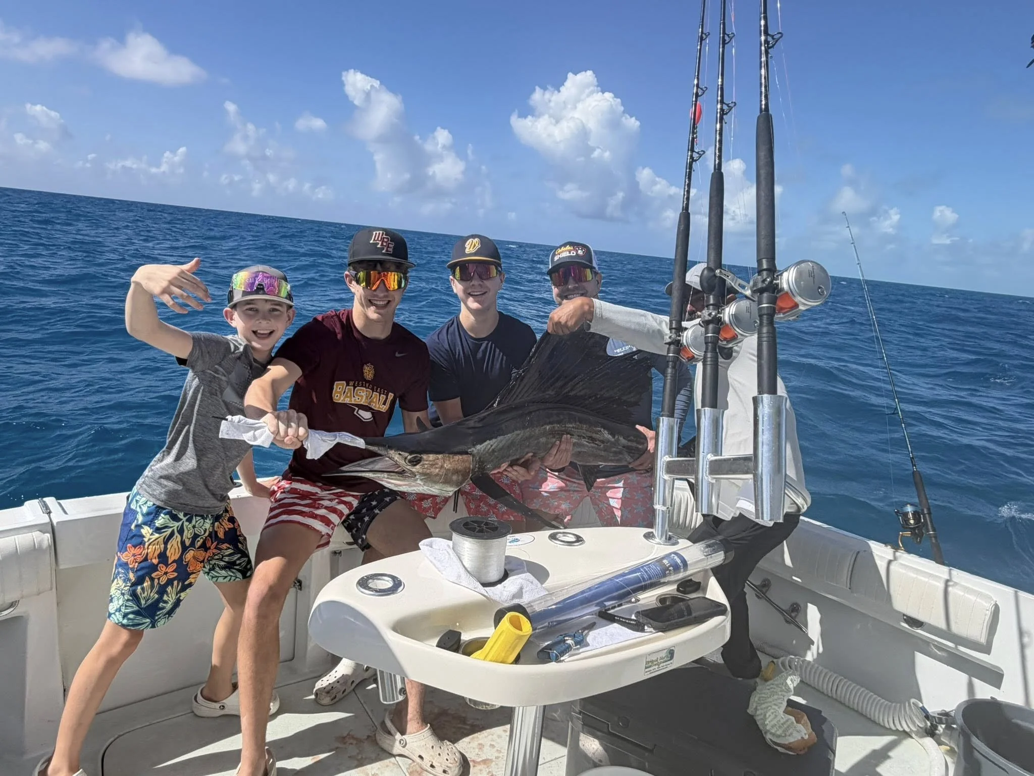Four people on a boat holding a large fish they caught, with fishing rods and the open ocean in the background.