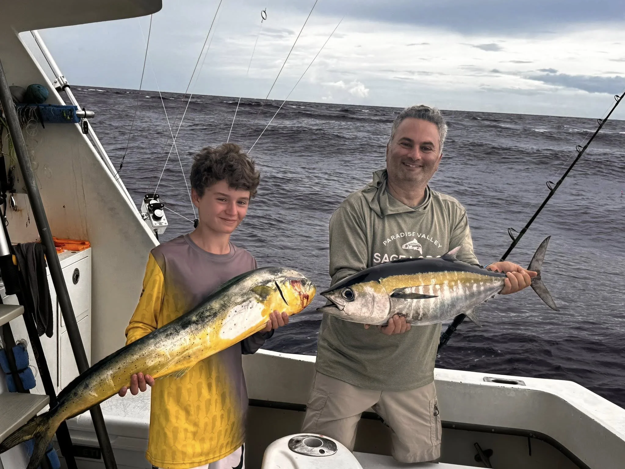 A man and a boy standing on a boat holding large fish they caught, with the ocean and cloudy sky in the background.