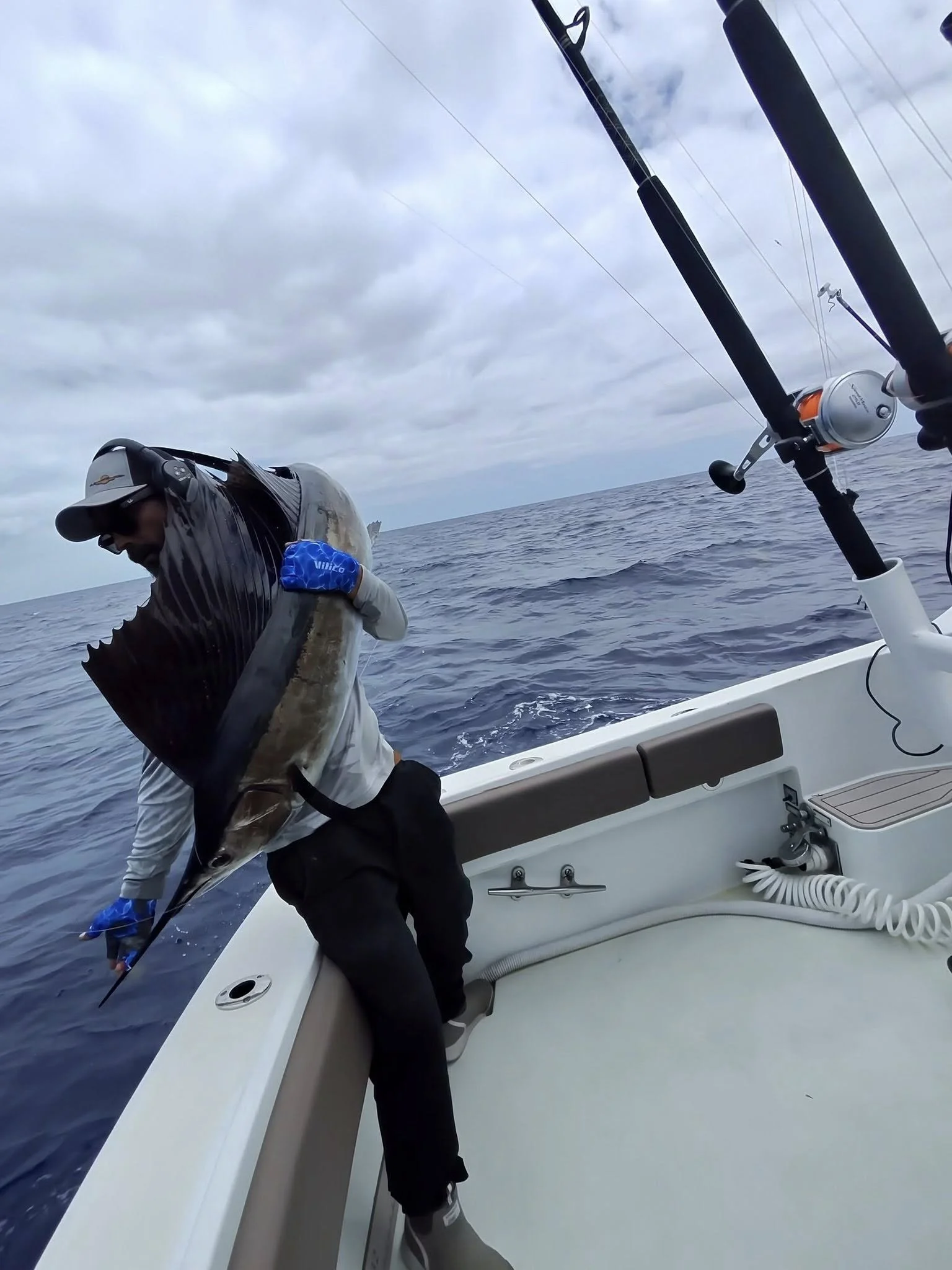 A person on a boat holding a large fish with the ocean and cloudy sky in the background, fishing rods visible.