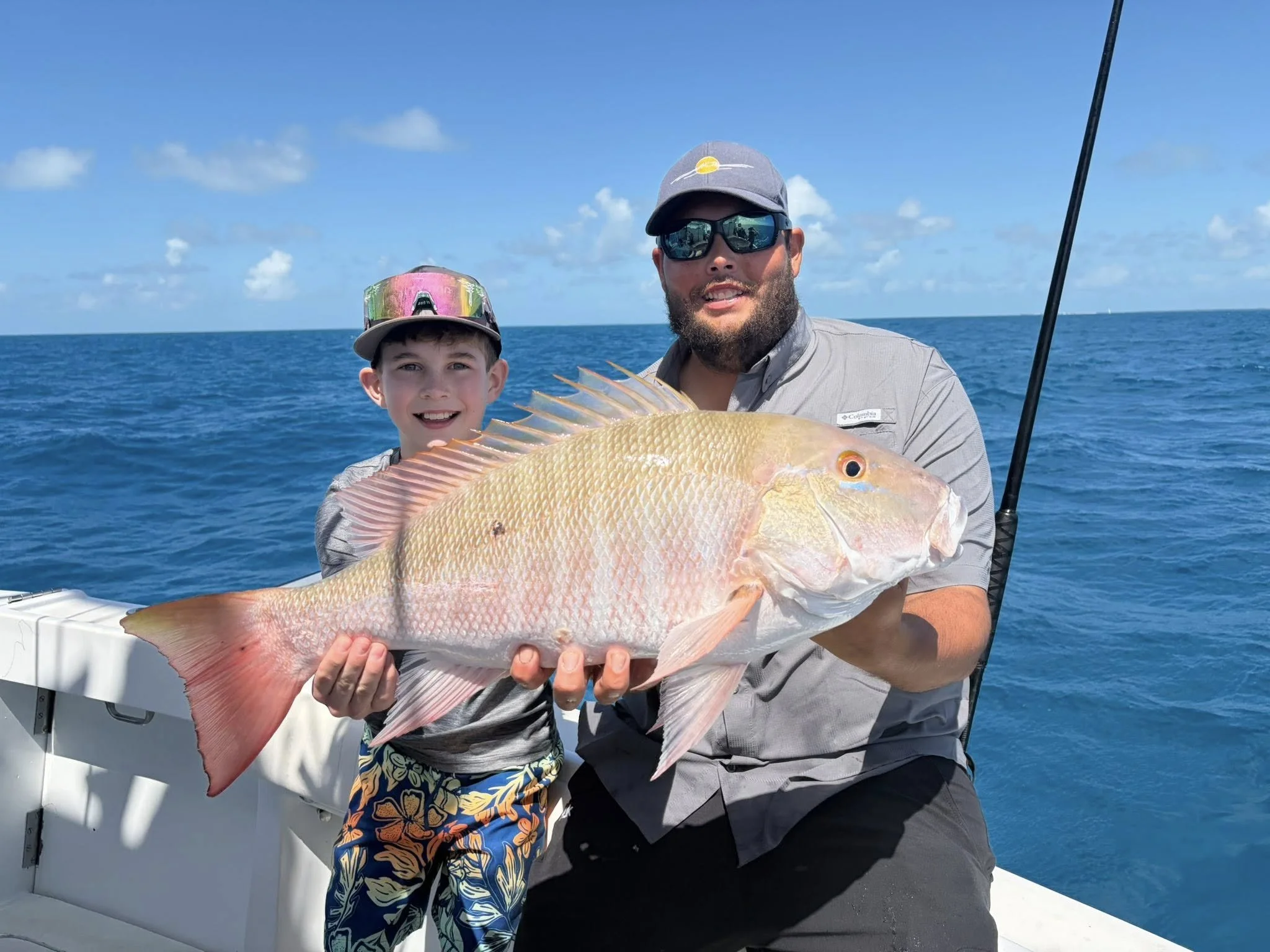 A man and a boy on a boat holding a large orange and pink fish with the ocean and blue sky in the background.