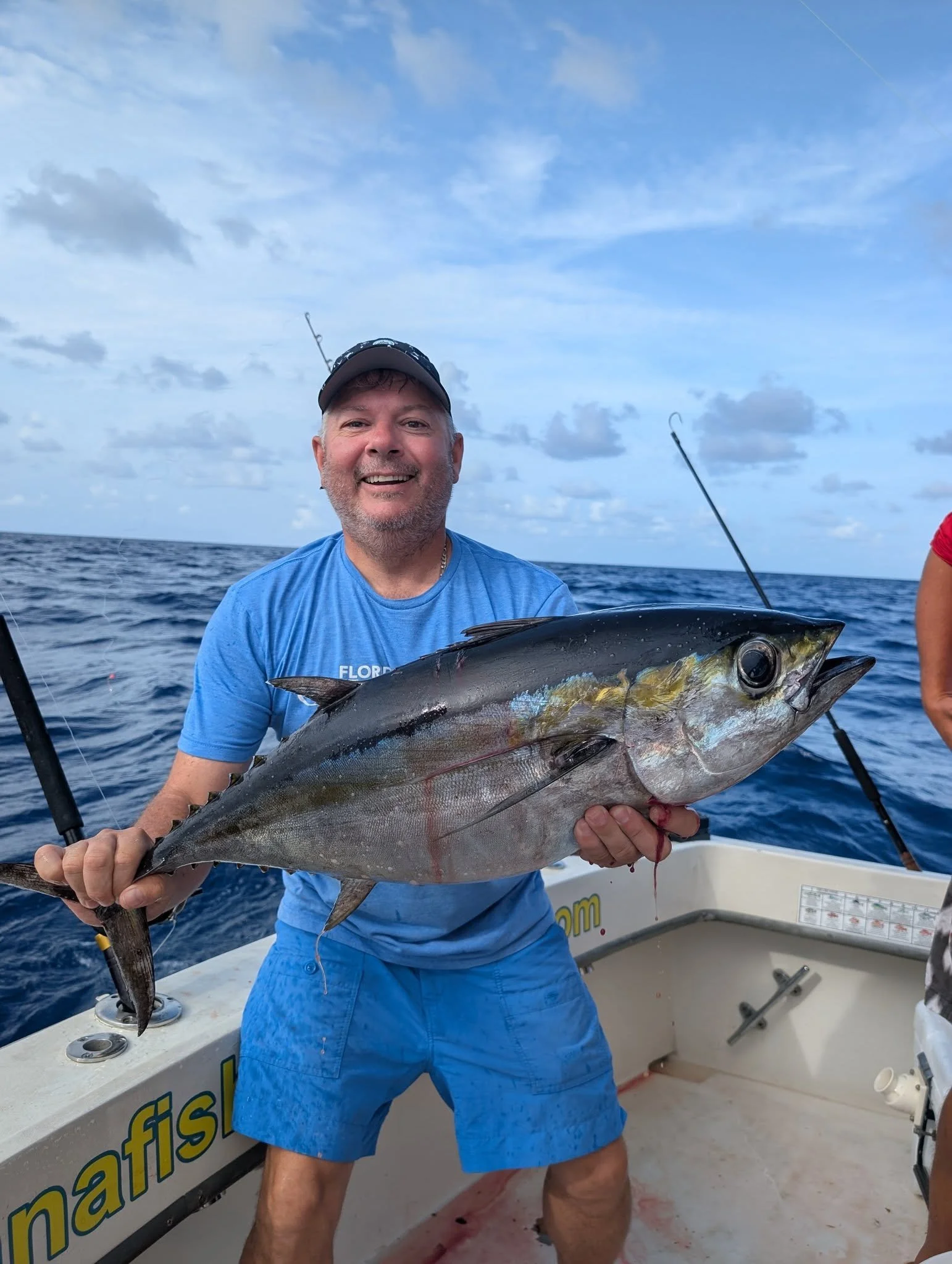 Man smiling and holding a large fish on a boat at sea, with fishing rods visible in the background under a partly cloudy sky.