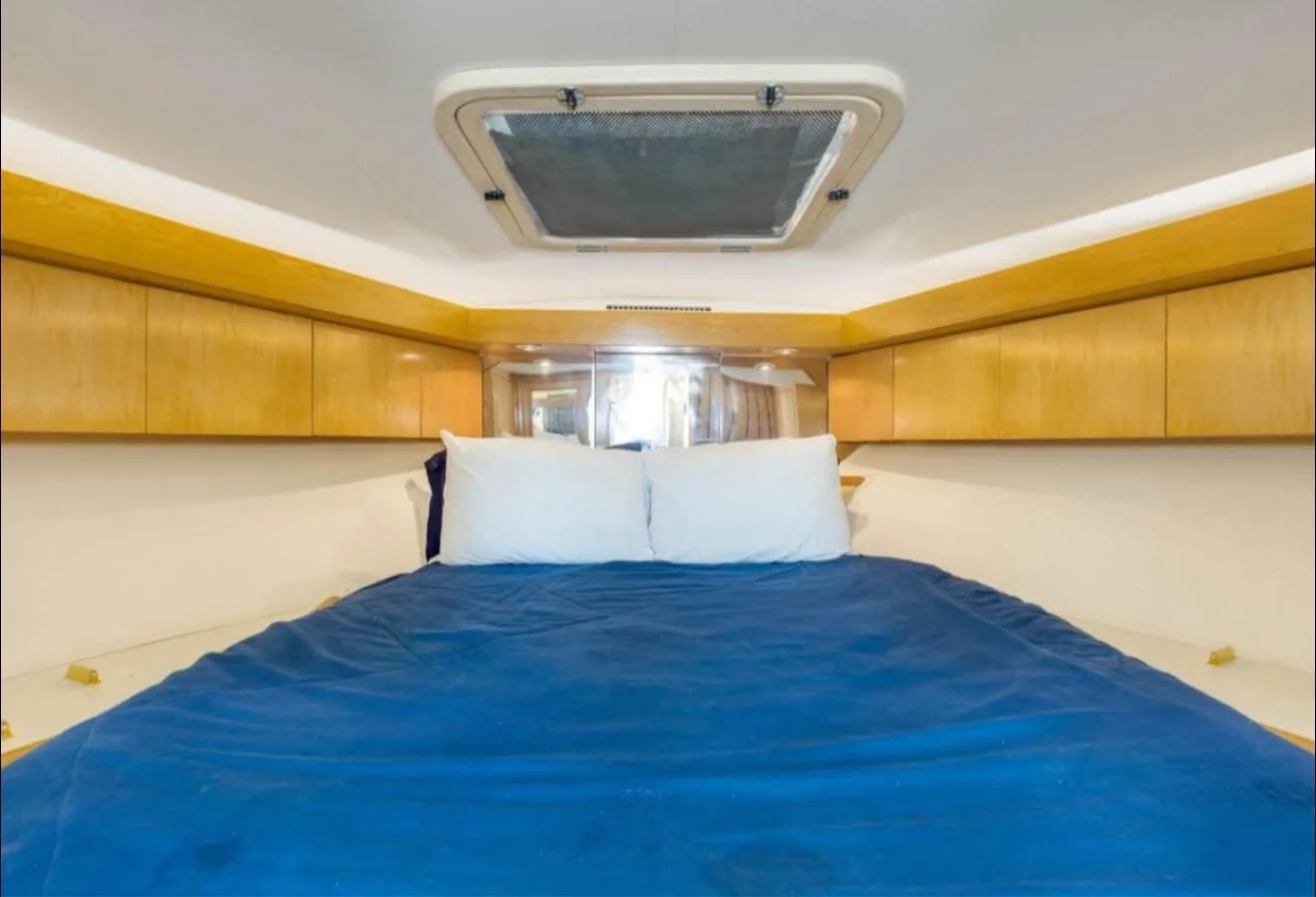View of a bed with white pillows and a blue blanket inside a boat cabin with wooden panels and overhead storage compartments.