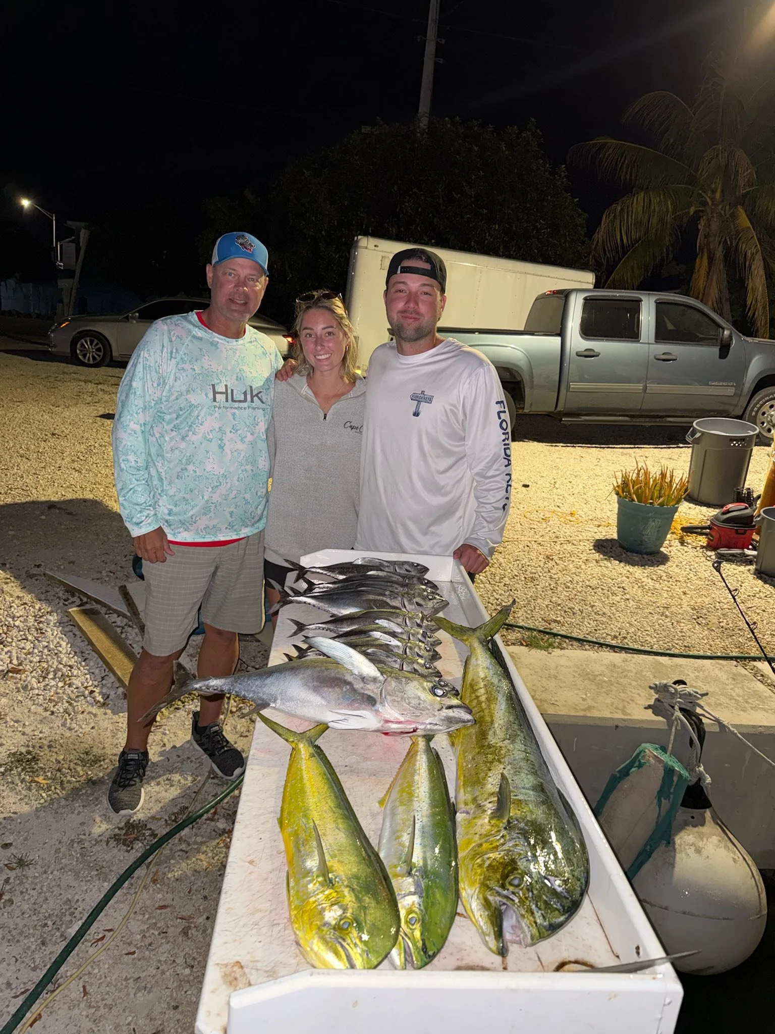 Three people standing behind a table with a variety of freshly caught fish, outdoors at night. There are cars and a palm tree in the background.