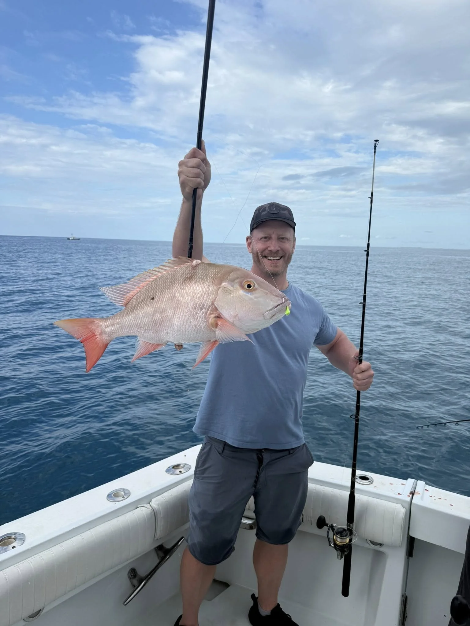 Man on a boat holding a large fish he caught, smiling at the camera, with a fishing rod in his other hand, in the ocean under a partly cloudy sky.