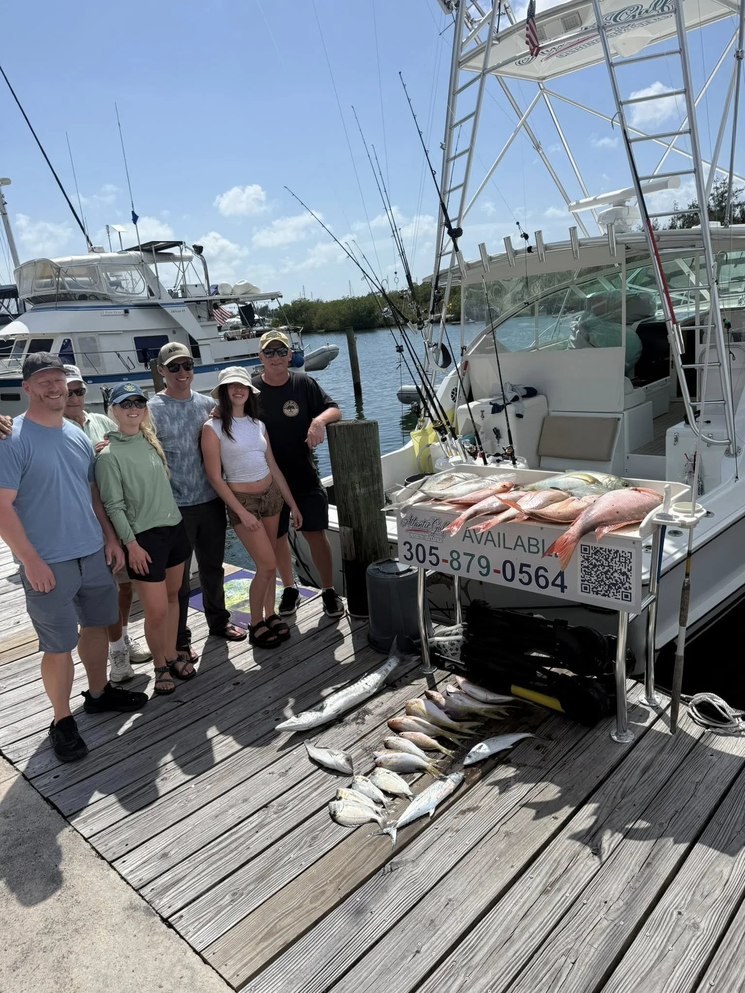 A group of six people standing on a dock next to a boat with caught fish displayed on a table and the dock. The boat has fishing rods mounted on it. The background shows more boats in the water and a blue sky with clouds.