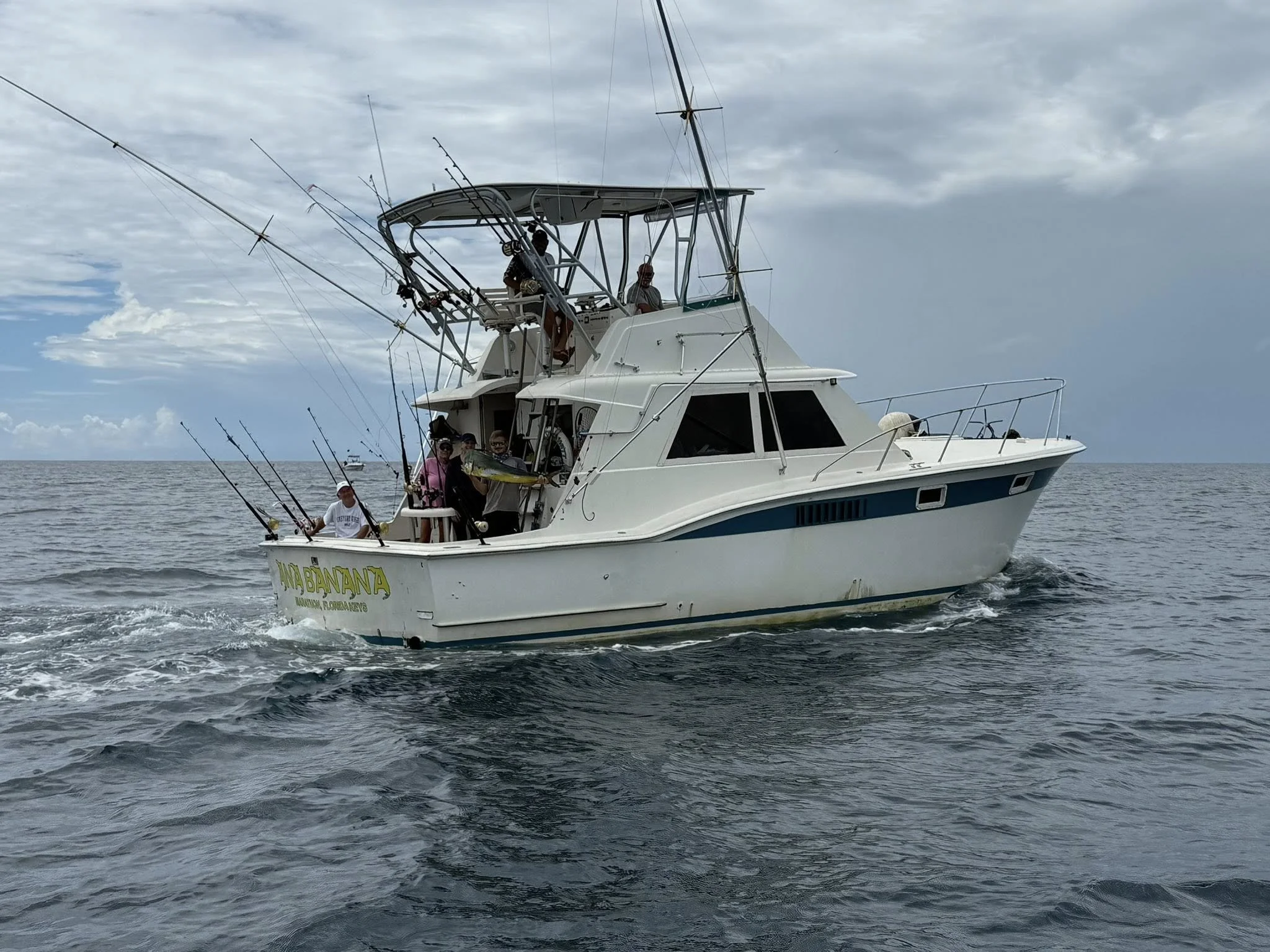 A white fishing boat named 'YNA BANANA' sailing on the ocean with several people on board, some fishing poles are attached to the boat, and the sky is partly cloudy.
