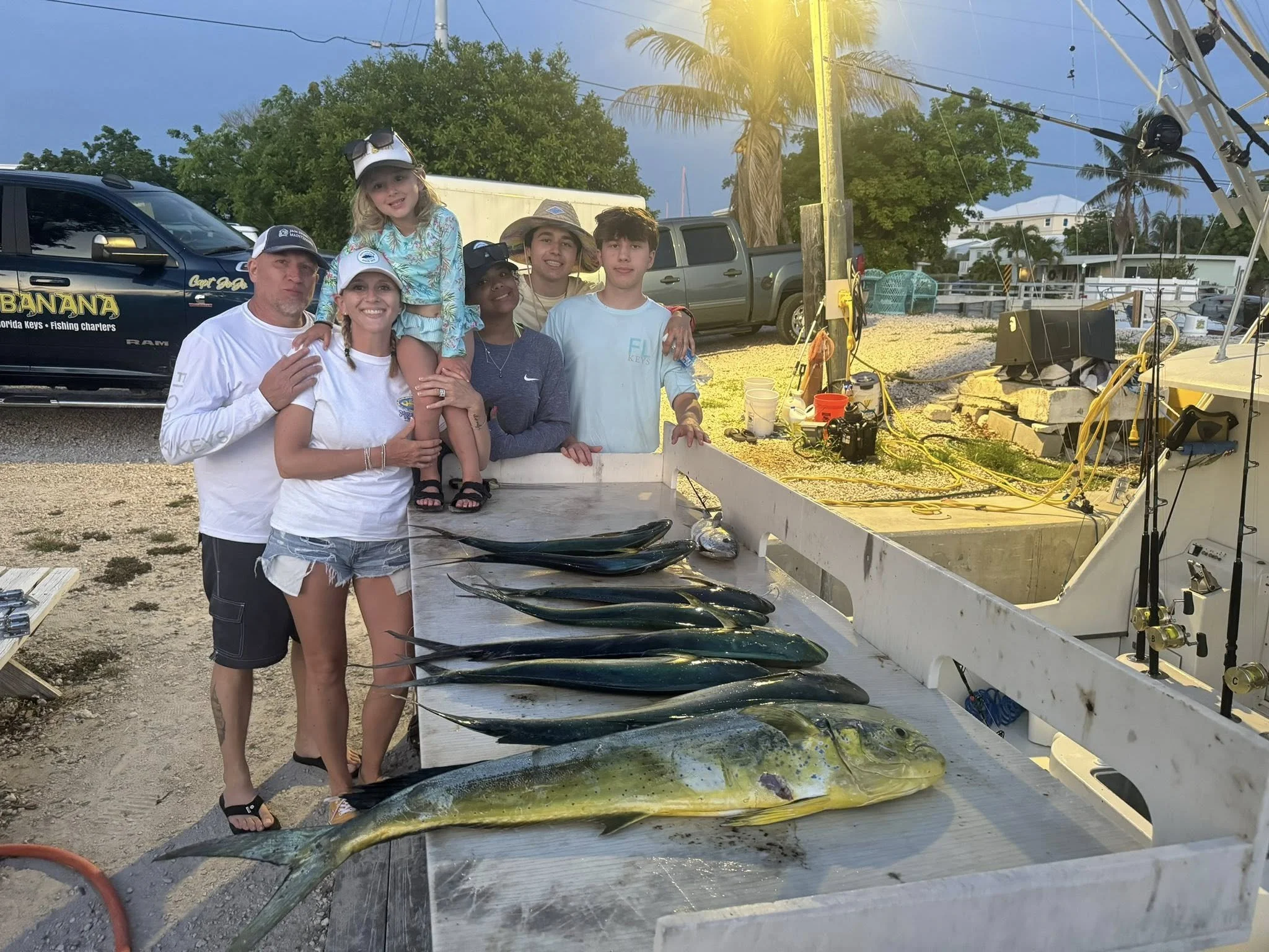 Family and friends standing behind a table with freshly caught fish at a dockside fishing spot during sunset, with boats and palm trees in the background.