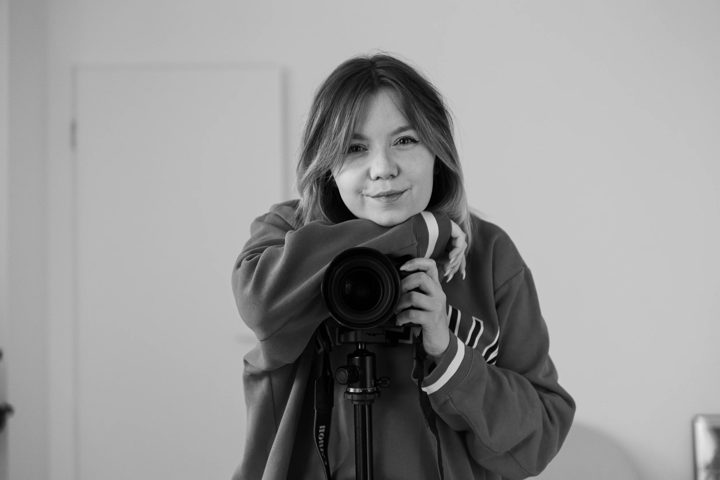 Schwarz-weiße Fotografie einer jungen Frau, die eine Kamera auf einem Stativ hält und in die Linse blickt.