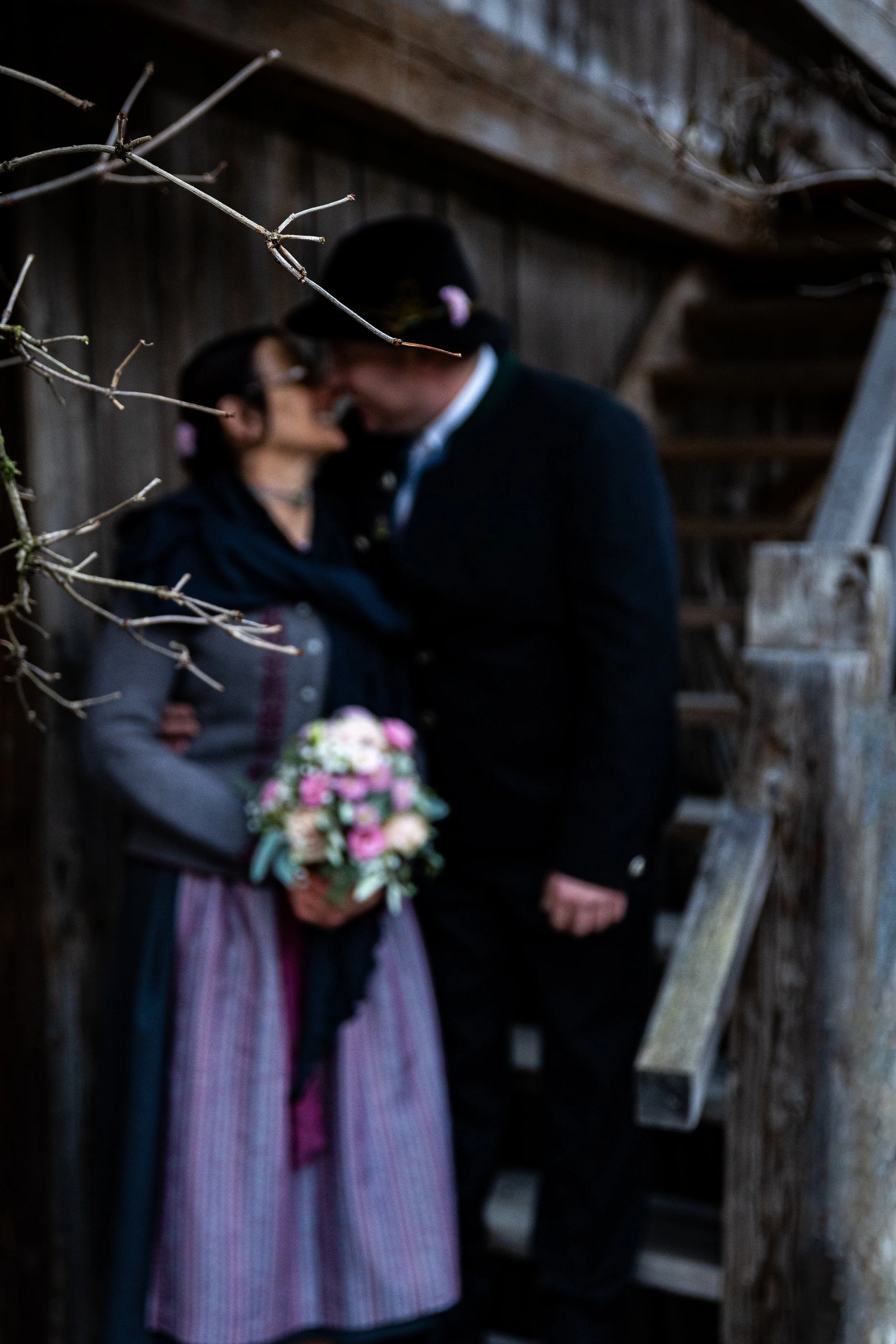 Ein Mann und eine Frau in traditionellen Trachten stehen sich nahe an einer Holztreppe und scheinen sich zu küssen. Die Frau hält einen Blumenstrauß in den Händen.