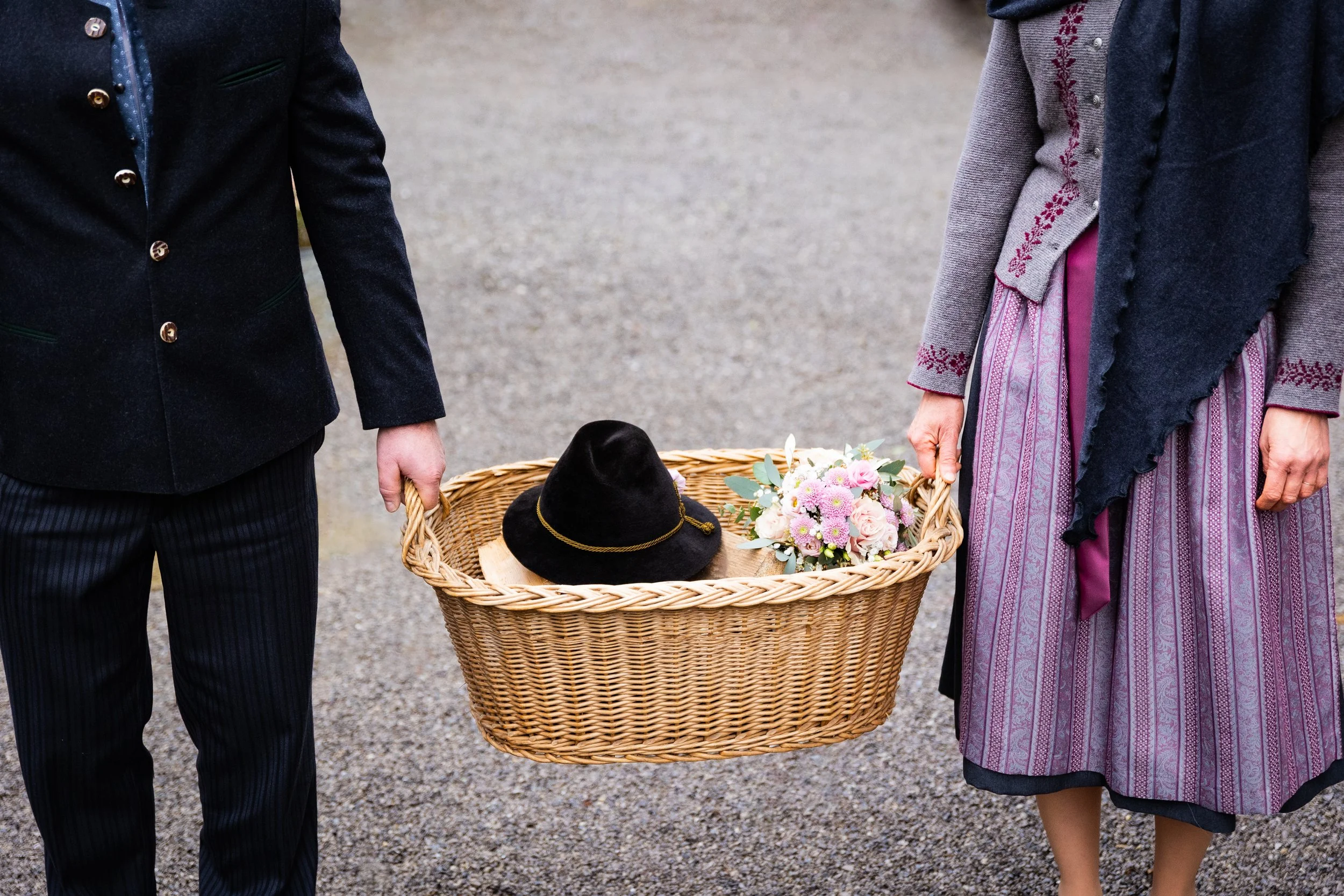Zwei Personen in traditionellen Trachten halten einen Korb mit einem schwarzen Hut und einem Blumenstrauß.