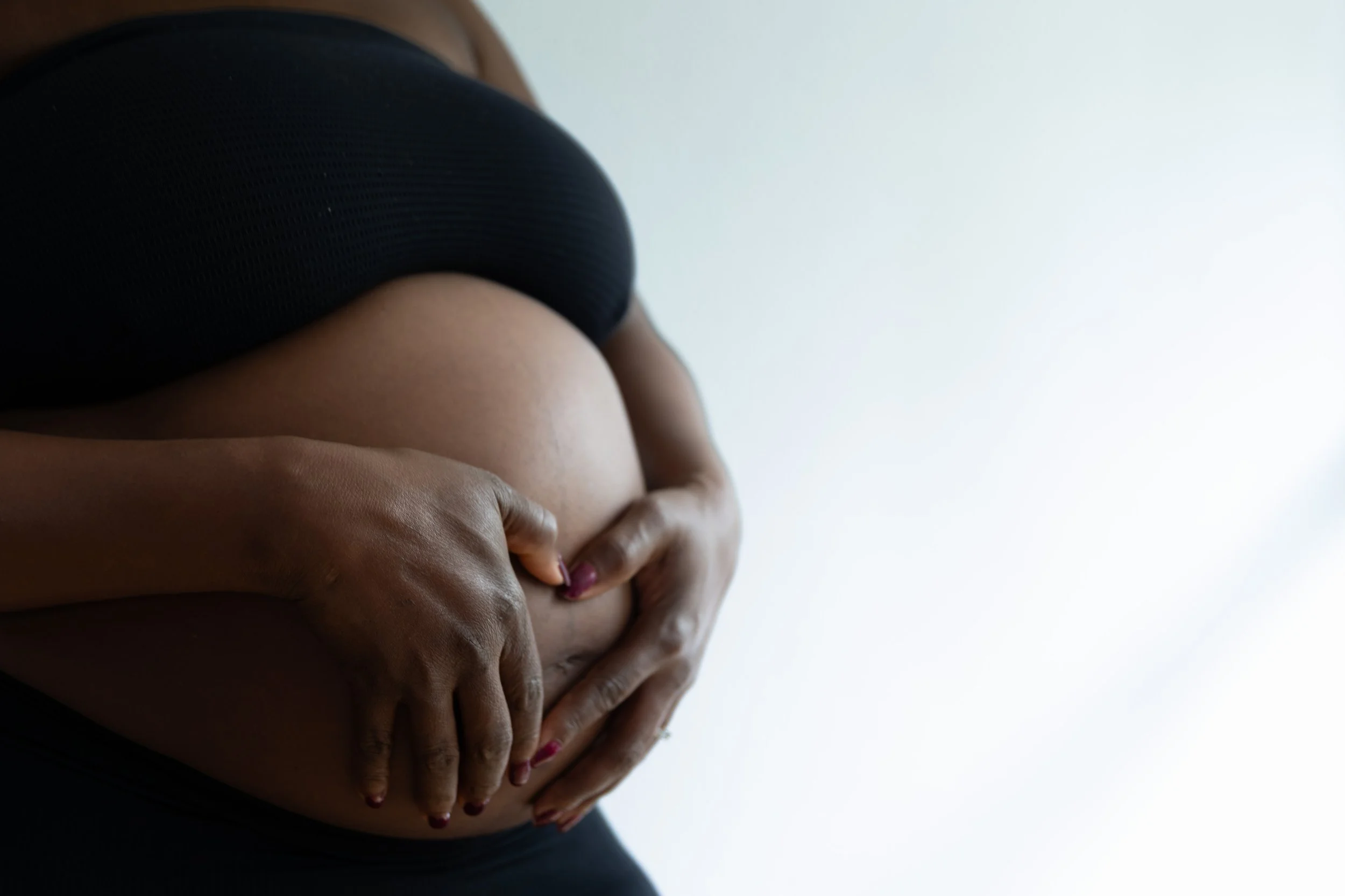 Schwangere Frau, die ihren Babybauch hält, mit dunkler Haut, trägt schwarze Sportkleidung, vor hellem Hintergrund.