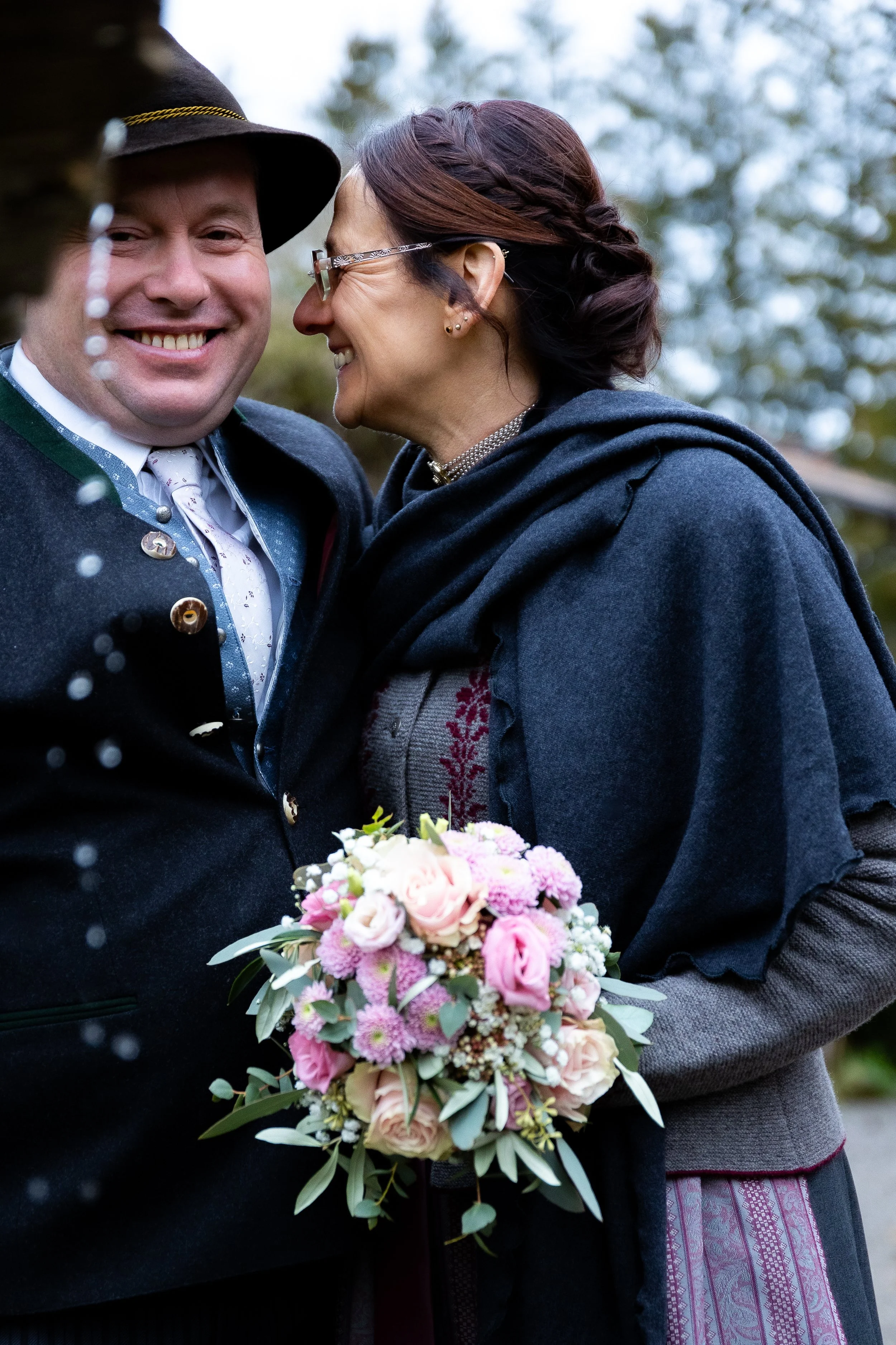 Ein glückliches Paar in traditioneller Kleidung bei einer Hochzeit, das sich küsst, mit einem Brautstrauß aus rosa und weißen Blumen.