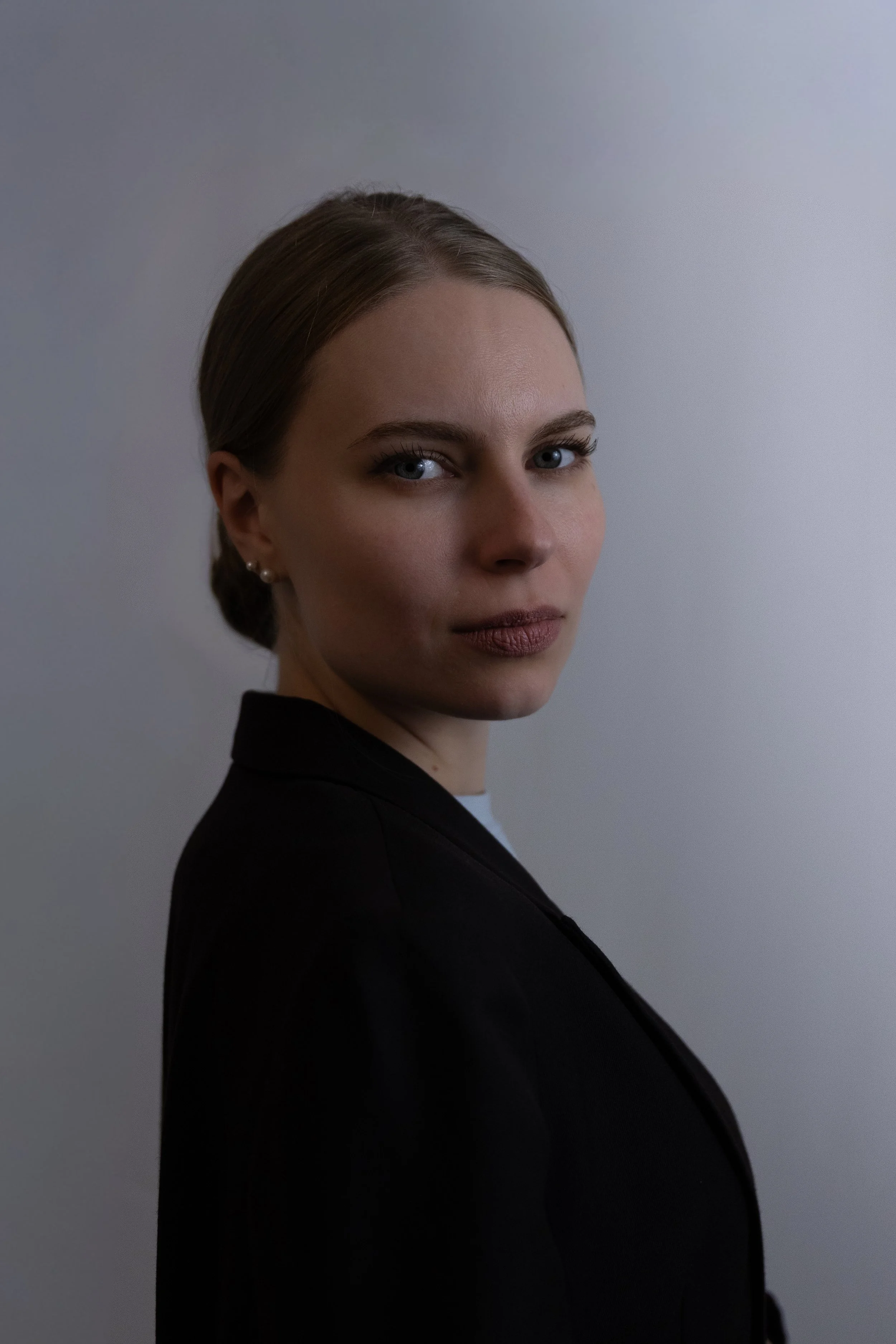 Porträt einer jungen Frau mit hellblauen Augen, braunen Haaren, dunklem Outfit und dezentem Make-up, vor neutralem grauem Hintergrund.