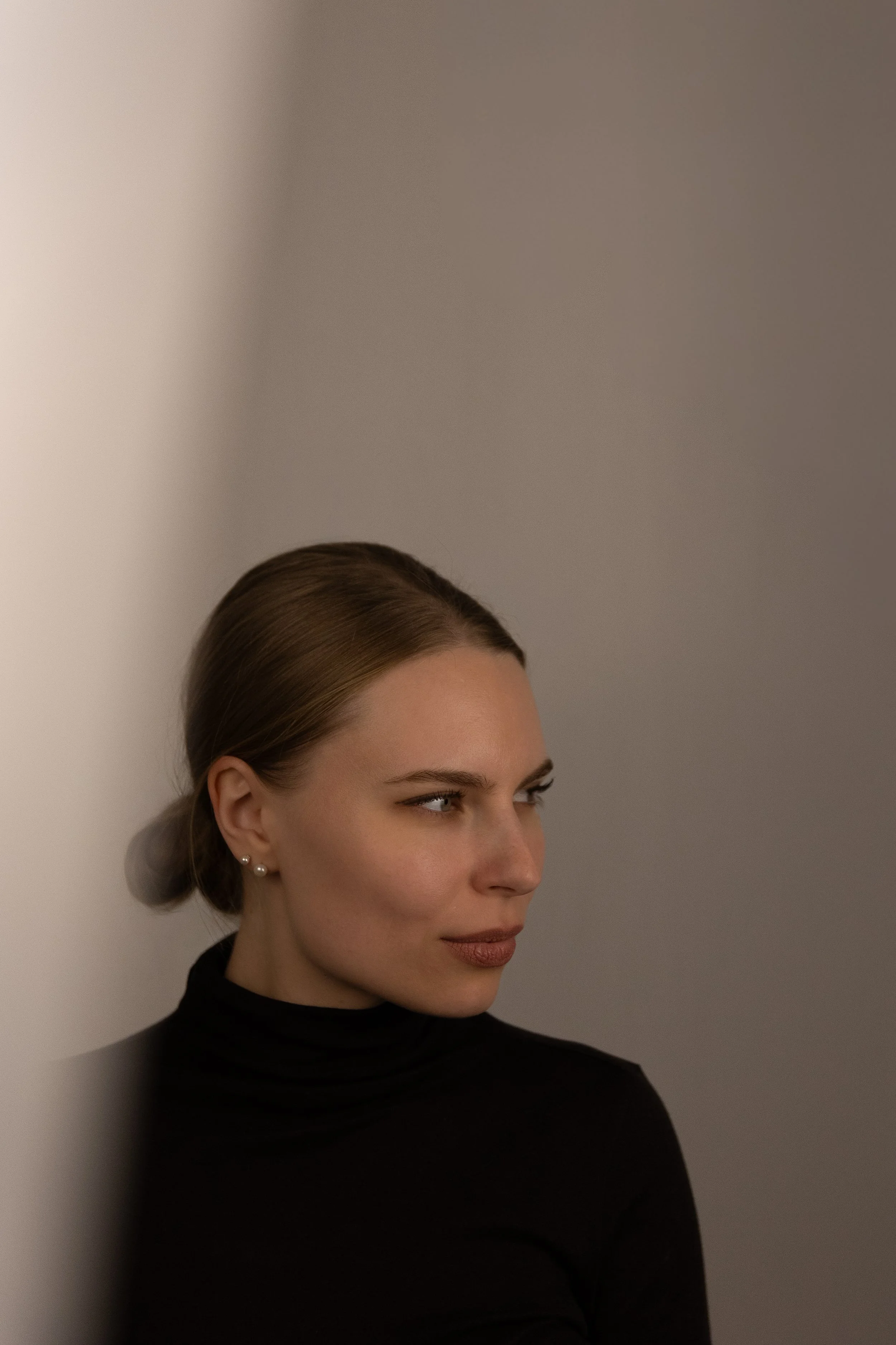 Porträt einer jungen Frau mit braunen Haaren, blauen Augen, hellen Hautfarbe, trägt Perlenohrringe und schwarzen Turtleneck, blickt seitlich nach rechts, vor einem neutralen, grauen Hintergrund.