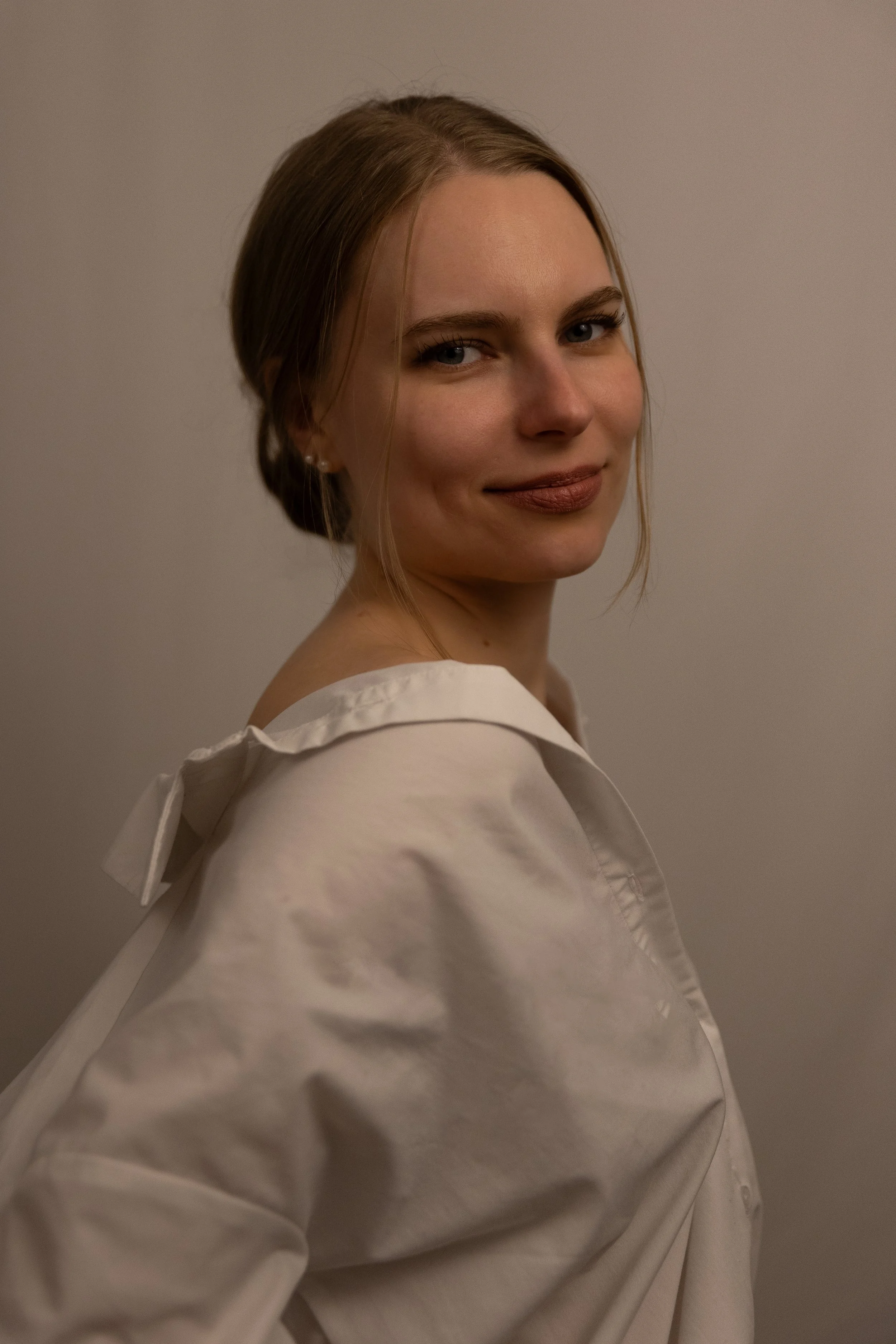 Das Bild zeigt eine junge Frau mit blonden Haaren, die seitlich lächelt, vor einem neutralen grauen Hintergrund. Sie trägt ein weißes Oberteil.