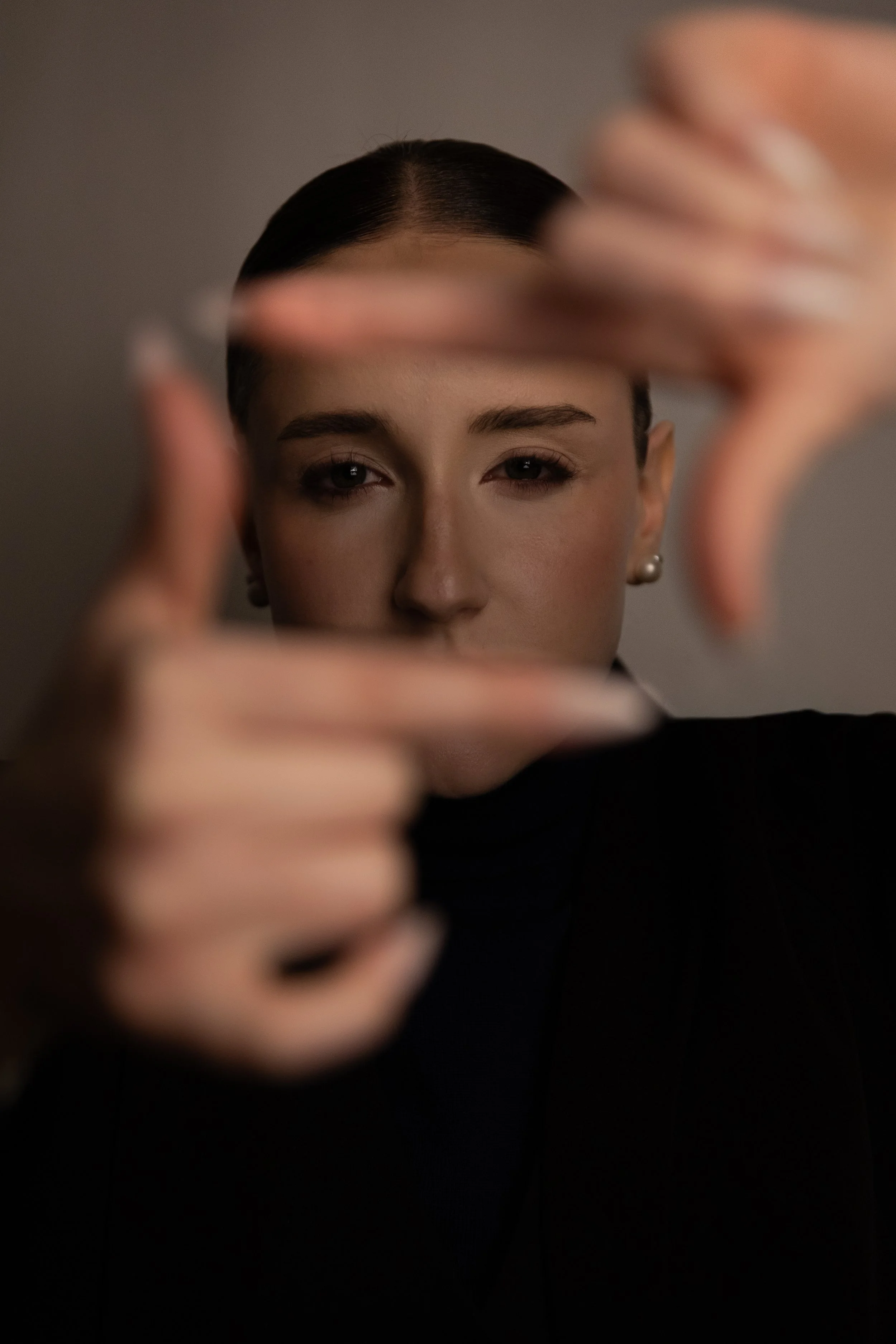Nahaufnahme eines gestylt Person mit kurzem, dunklem Haar, die durch eine Hand vor einer Kamera blickt.
