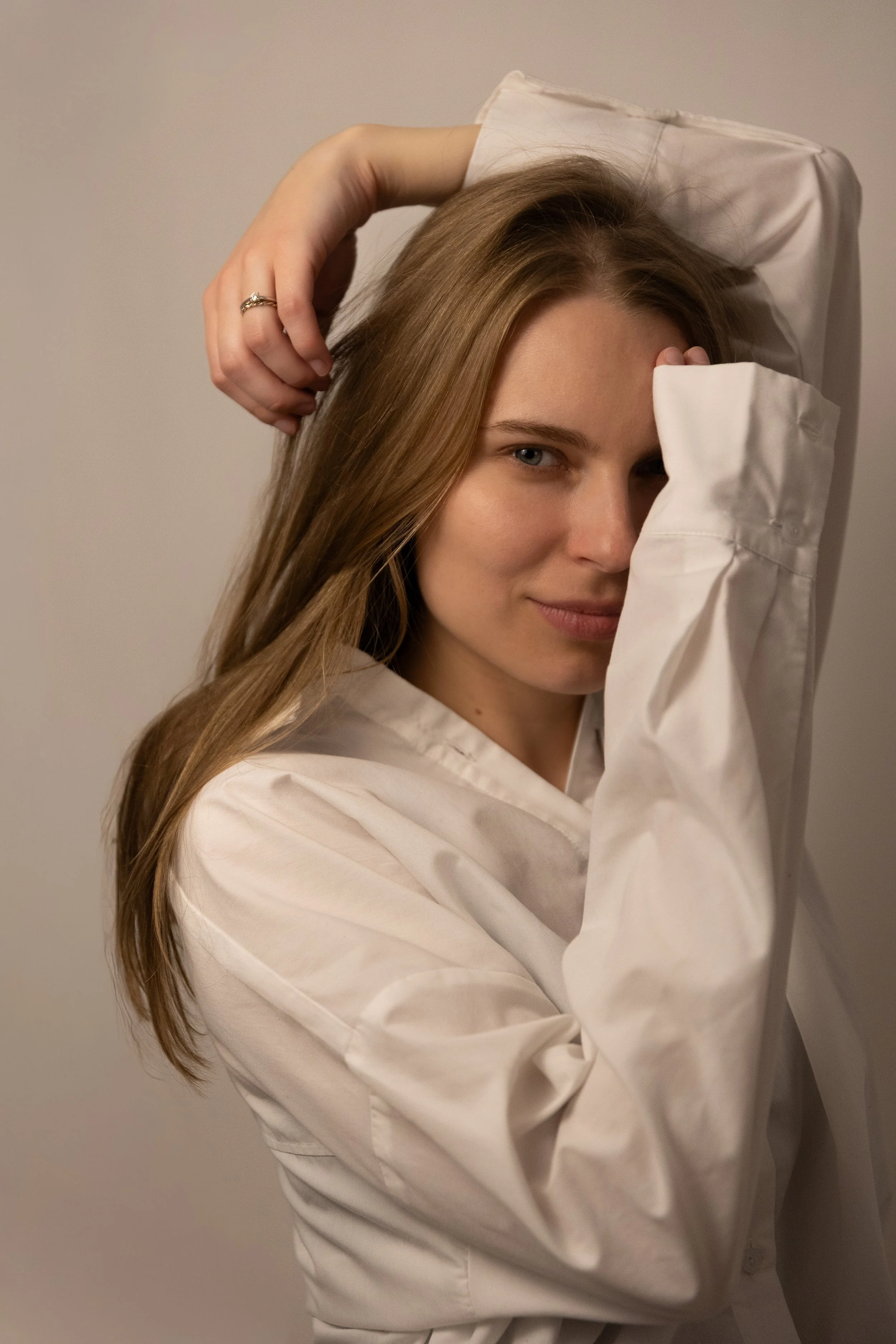 Junge Frau mit langen, braunen Haaren, trägt einen weißen Hemd, blickt schelmisch mit einem Lächeln, halb verdeckt ihr Gesicht mit einem Arm, steht vor neutralem Hintergrund.