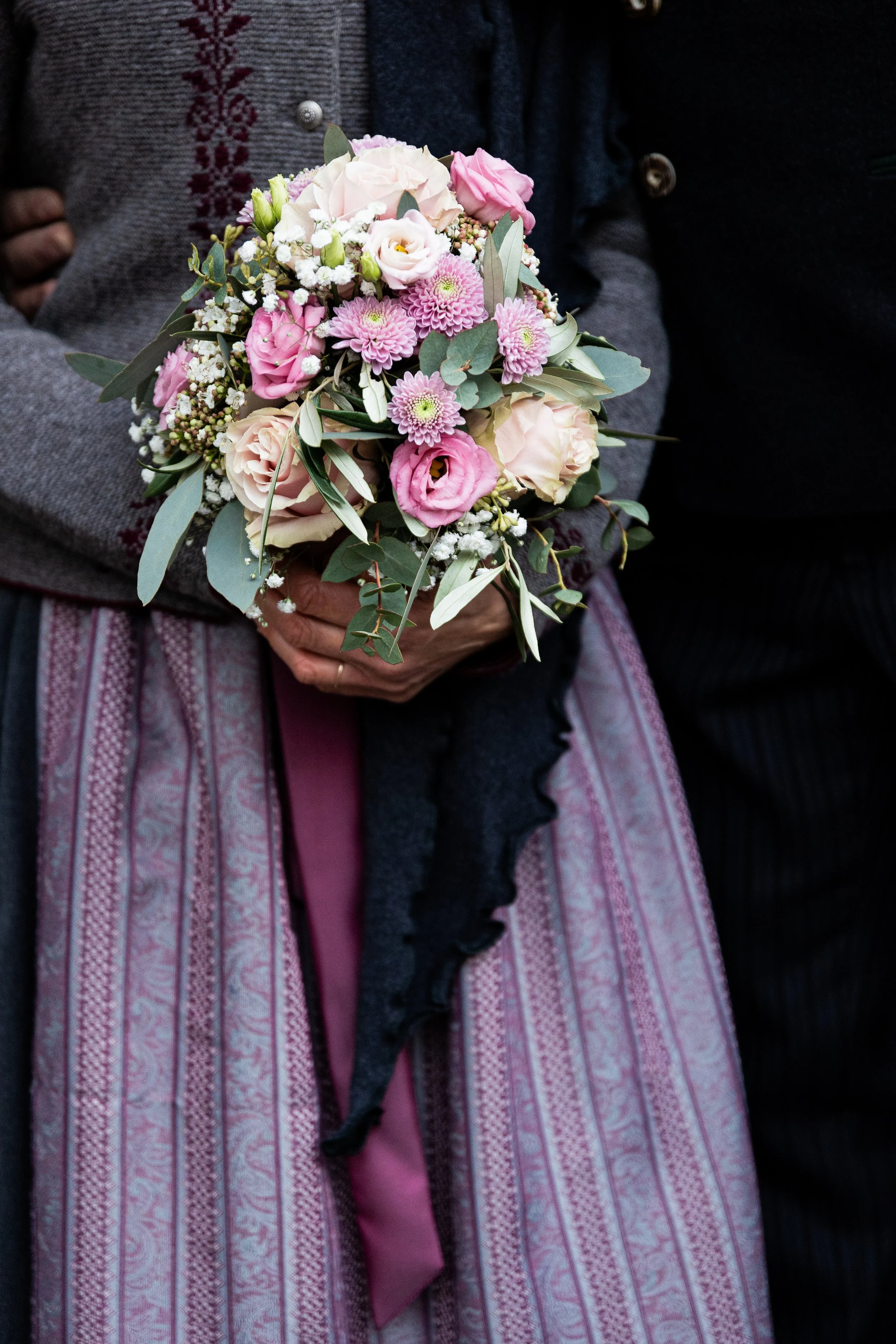 Eine Person hält einen Blumenstrauß mit rosa und weißen Blumen, umgeben von grünen Blättern. Die Person trägt ein traditionelles, geblümtes Kleid und ein Jackett.