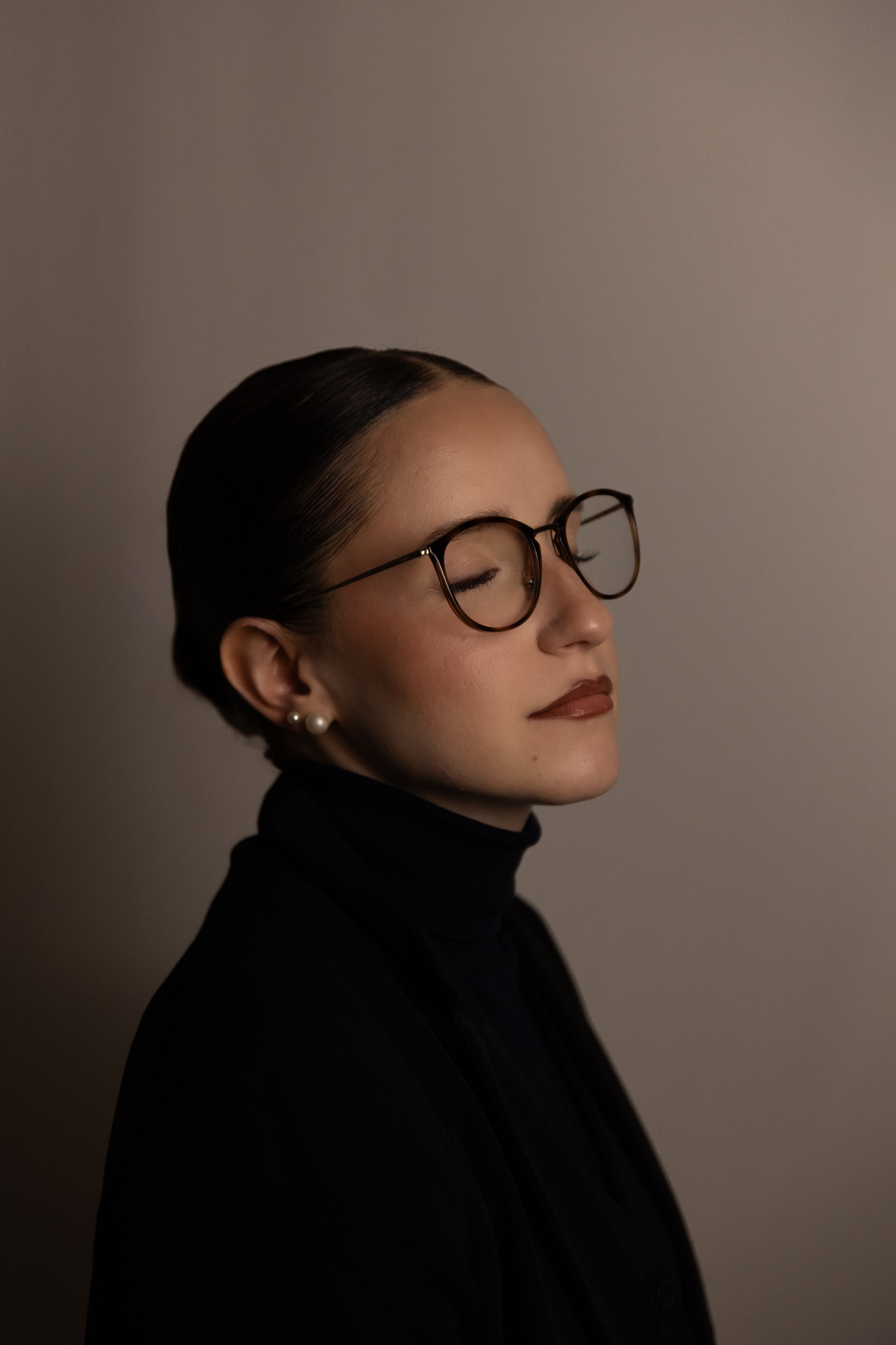 Porträt einer Frau mit dunklem Haar, Brille und Perlenohrringen, im Profil, vor einem neutralen grauen Hintergrund.