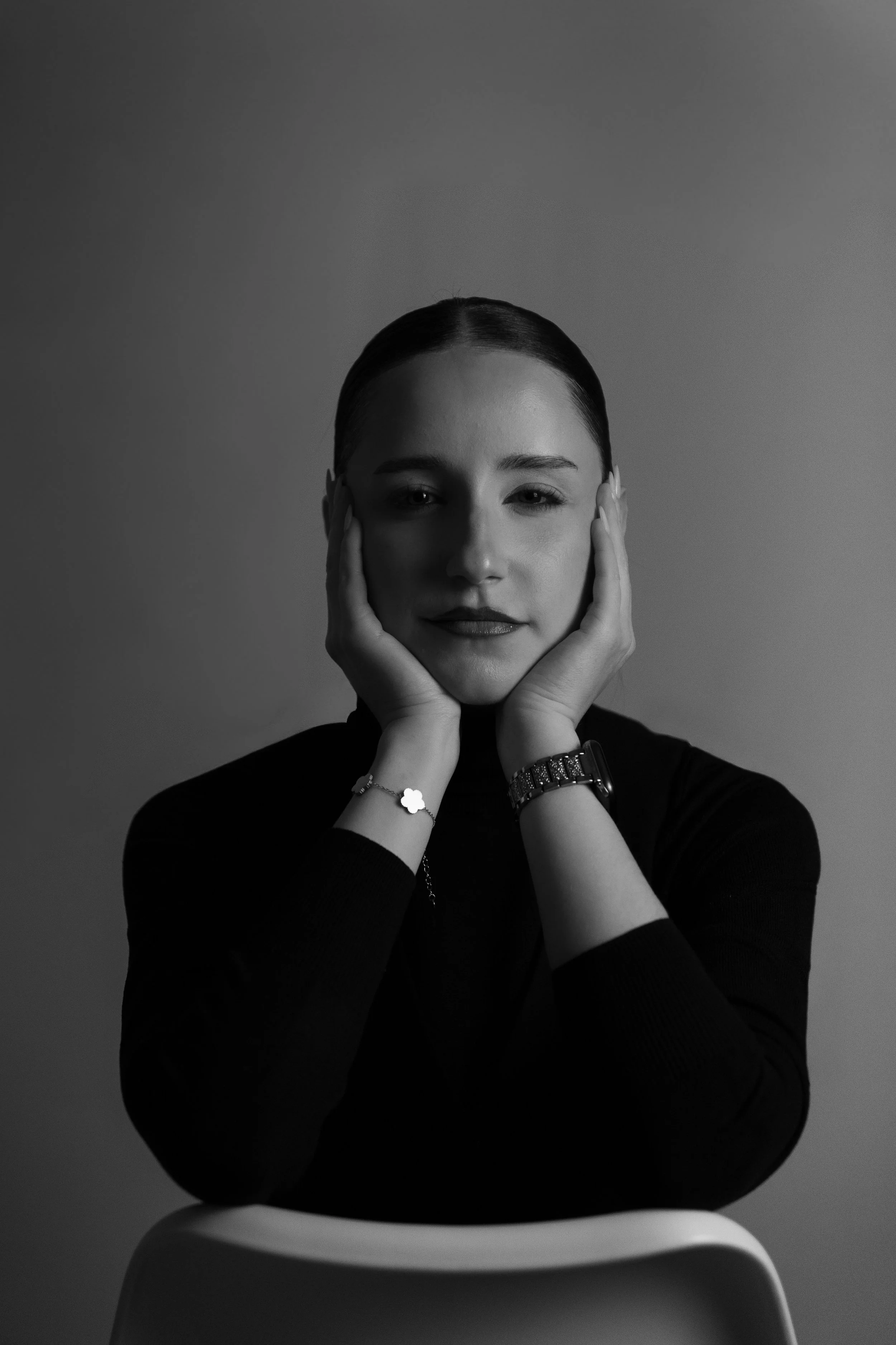 Schwarz-Weiß-Foto eines jungen Frauenporträts, die auf einem Stuhl sitzt, den Kopf mit Händen stützt und direkt in die Kamera schaut.