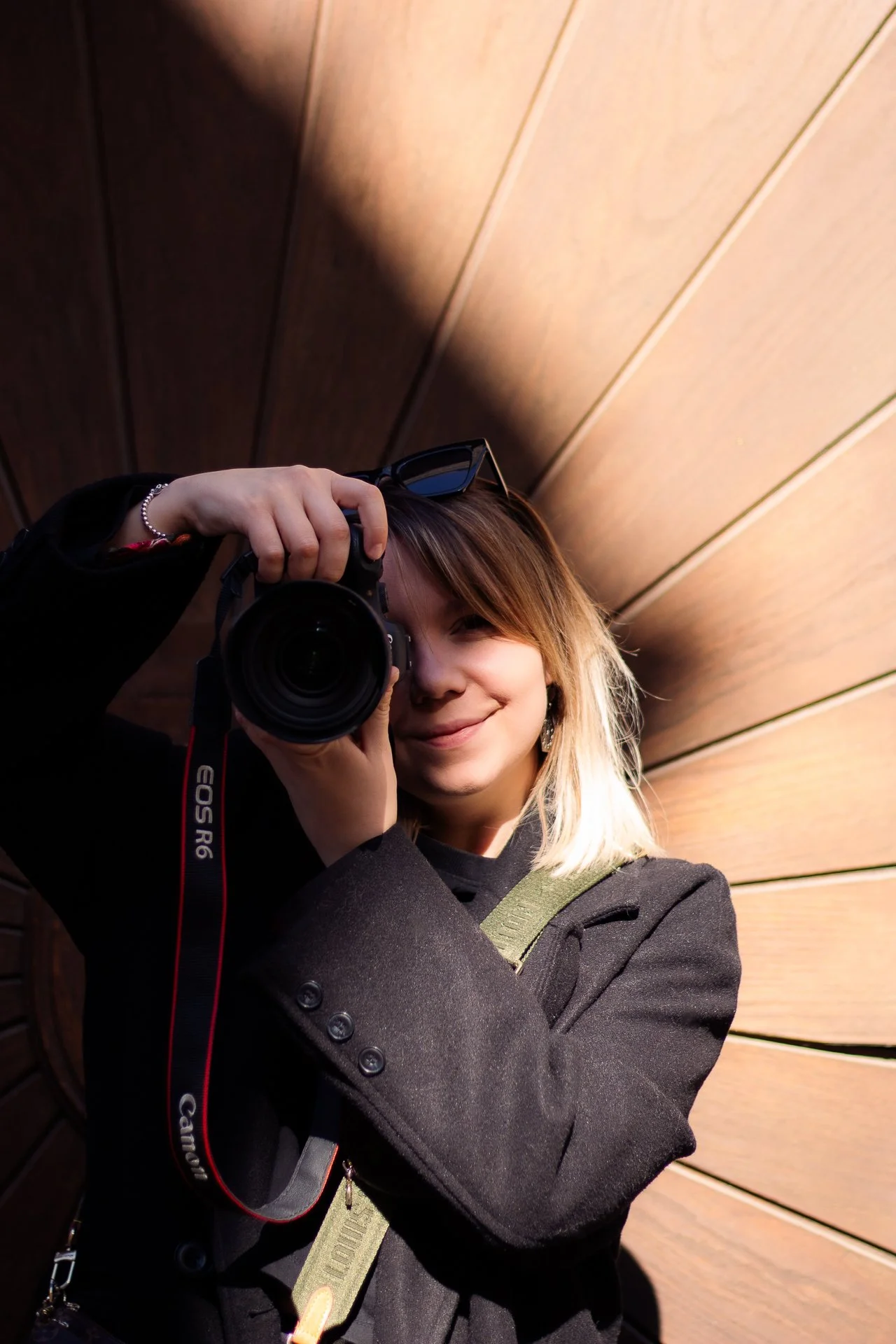 Junge Frau mit Kamera, die in die Linse schaut, vor einer Holzwand, Fotografenbrille auf dem Kopf, lächelnd.