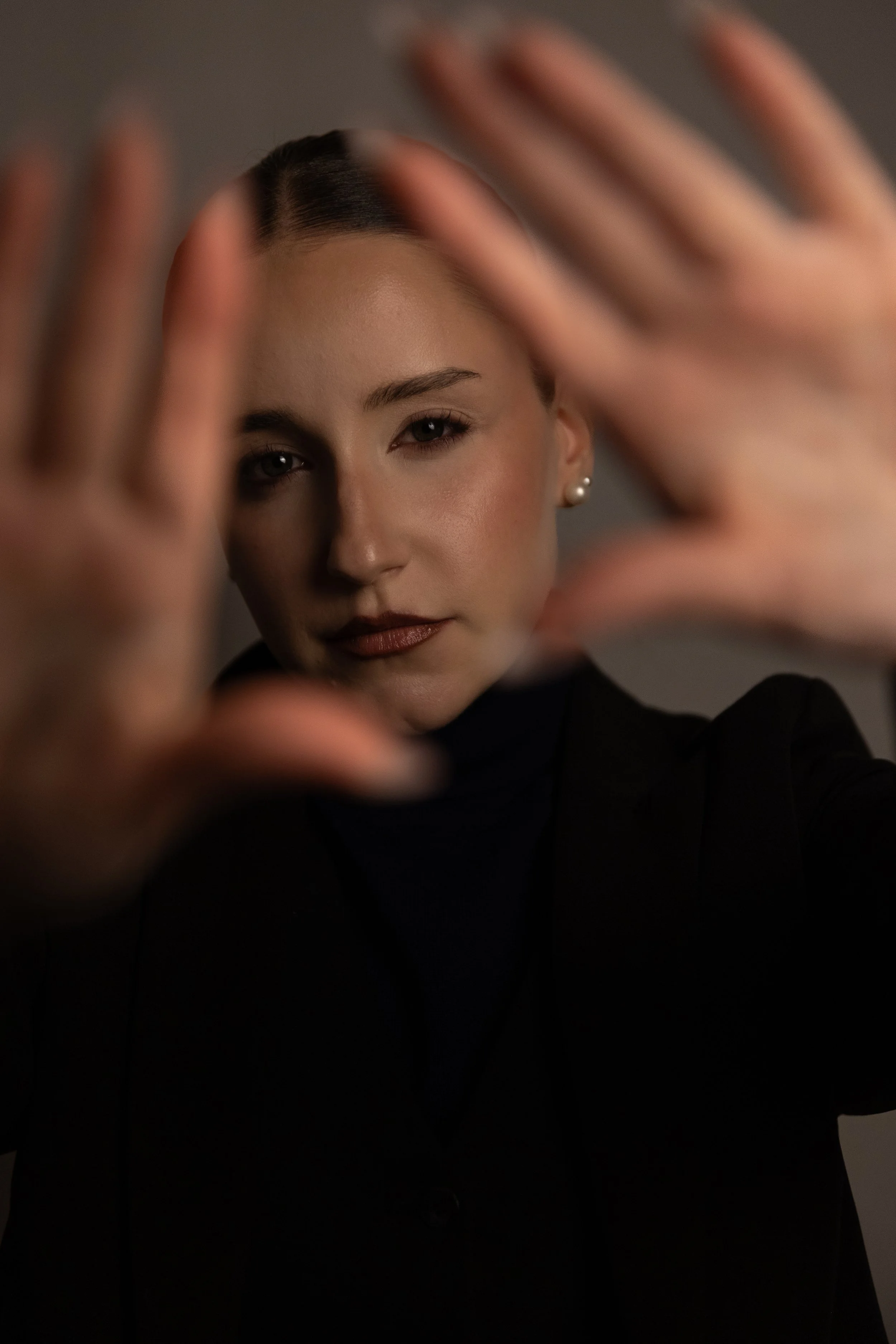 Porträt einer jungen Frau mit dunklem Haar, die ihre Hände fast vollständig vor ihr Gesicht hält, wobei nur ein Teil ihres Gesichtes sichtbar ist. Sie trägt Ohrlöcher und hat einen selbstbewussten Gesichtsausdruck.