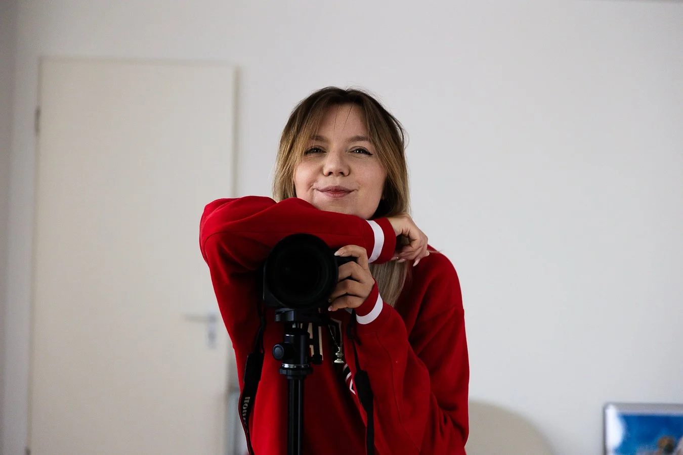 Junge Frau macht Selfie mit Kamera, trägt rotes Sweatshirt, steht vor weißer Wand in einem Raum.
