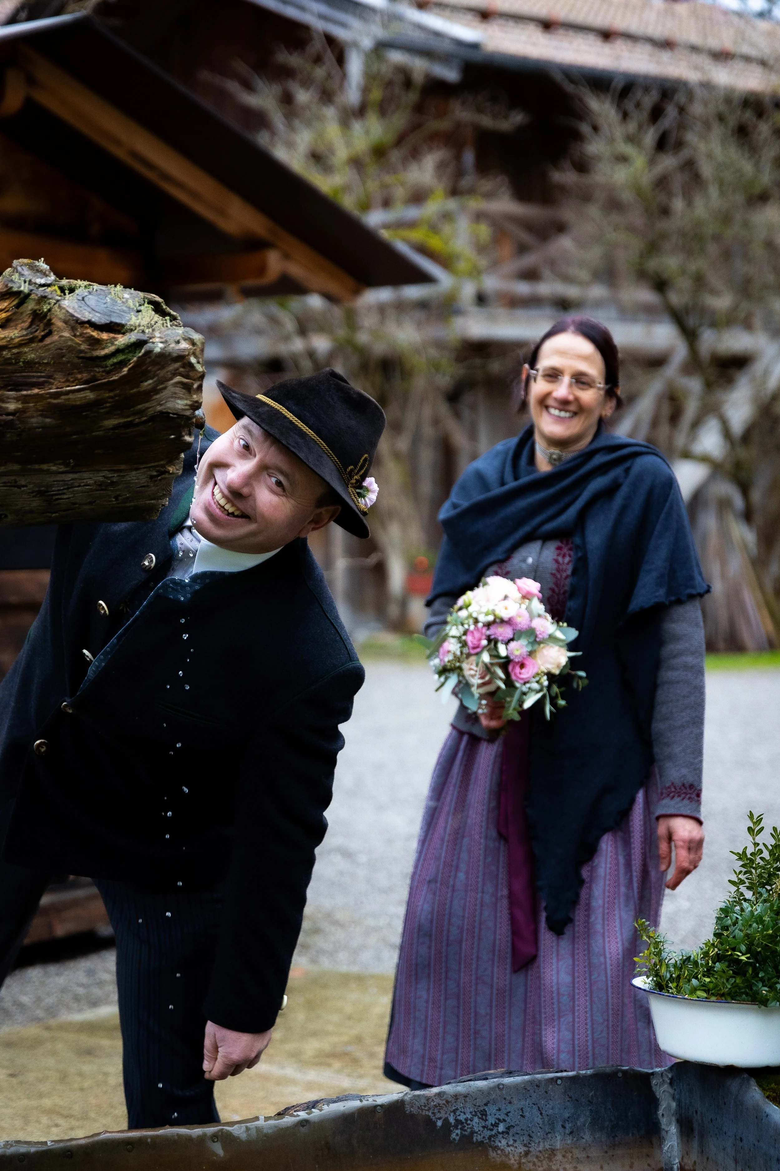 Ein Mann in traditioneller Tracht lächelt in die Kamera, während eine Frau im Vintage-Stil einen Blumenstrauß hält und beide vor einem holzverschindelten Gebäude stehen.