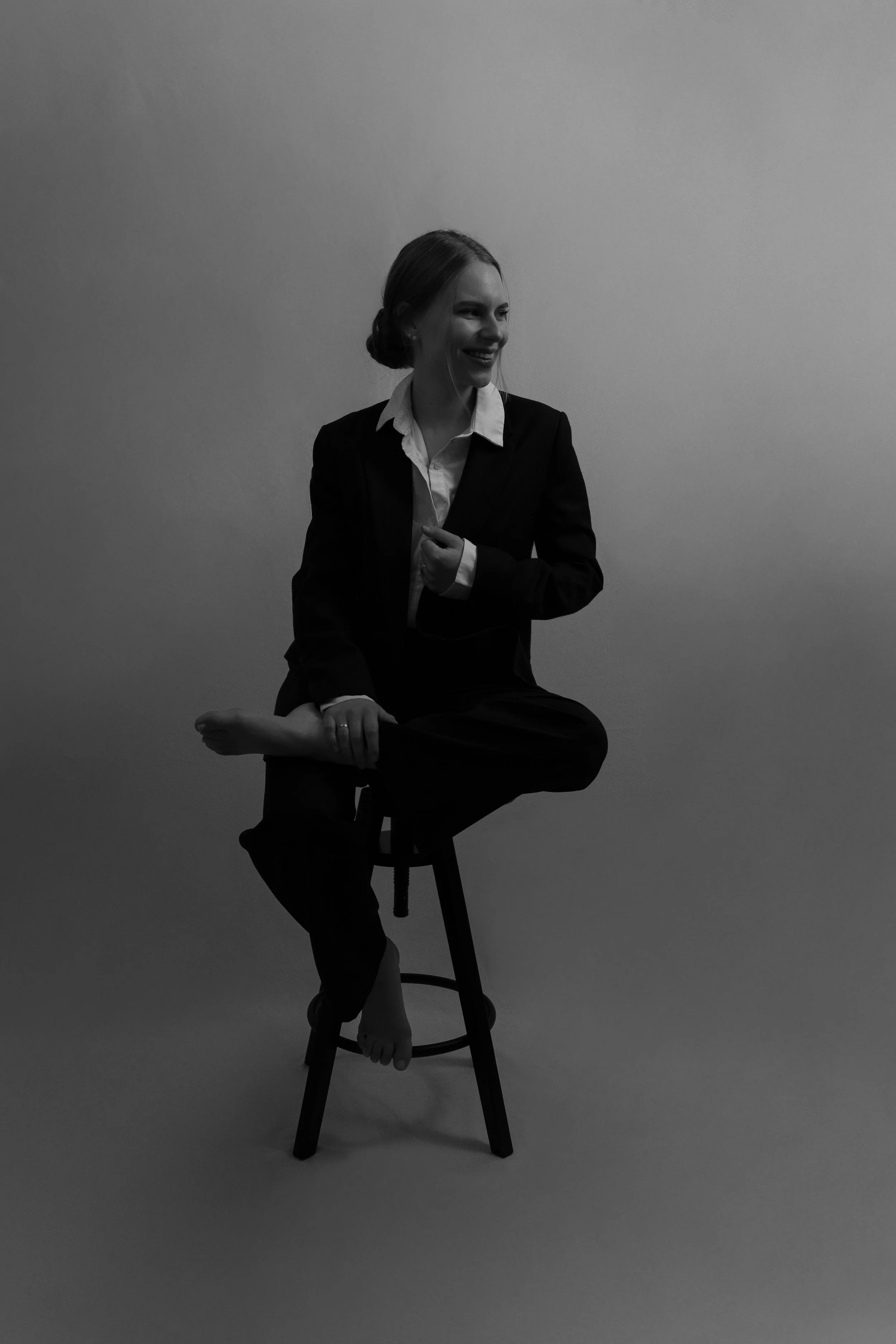 Schwarz-weiß-Foto einer lachenden Frau in formeller Kleidung, die auf einem Hocker sitzt, mit einem minimalistischen, hellen Hintergrund.